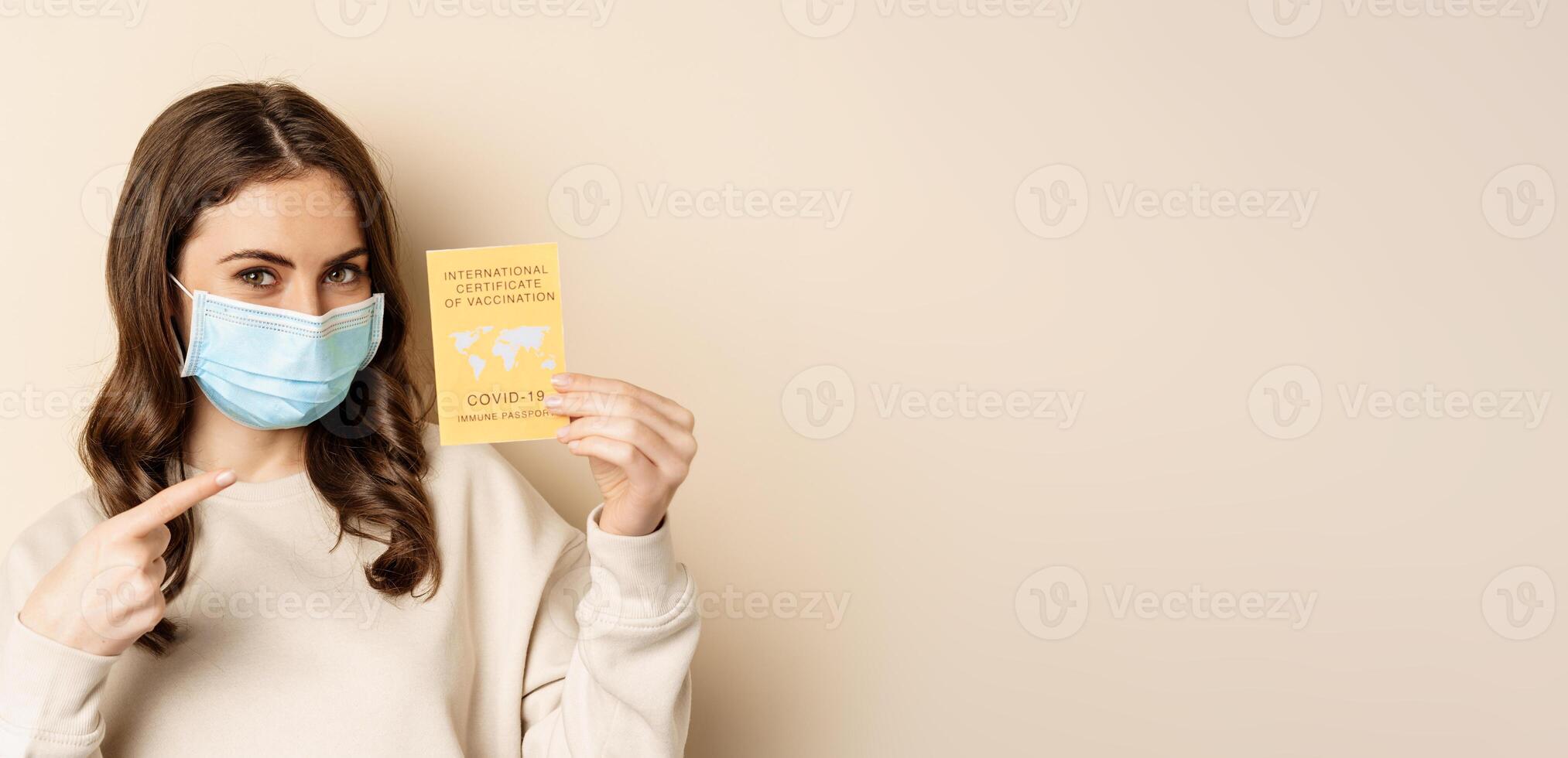 Covid and people concept. Vaccinated woman in face mask showing covid international vaccination certificate, recommend get vaccine, standing over beige background photo
