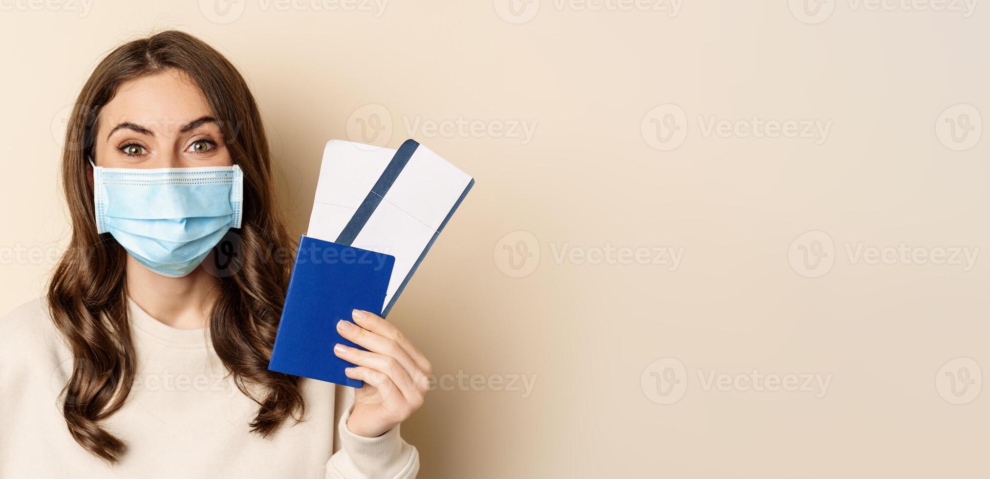 Travel and covid-19 pandemic. Close up portrait of smiling woman in medical mask, showing passport with two tickets, concept of tourism, beige background photo