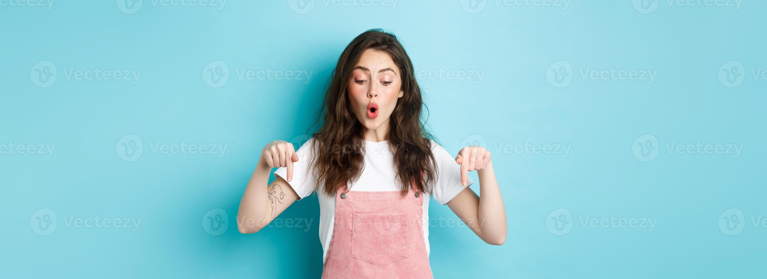 Amazed young beautiful woman checking out promo deal, pointing fingers down and looking fascinated, saying wow, standing over blue background photo