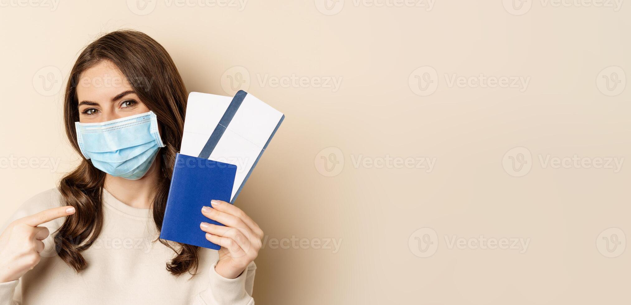 Travel and covid-19 pandemic. Girl in medical face mask travelling, showing passport with two tickets on plane, standing against beige background photo