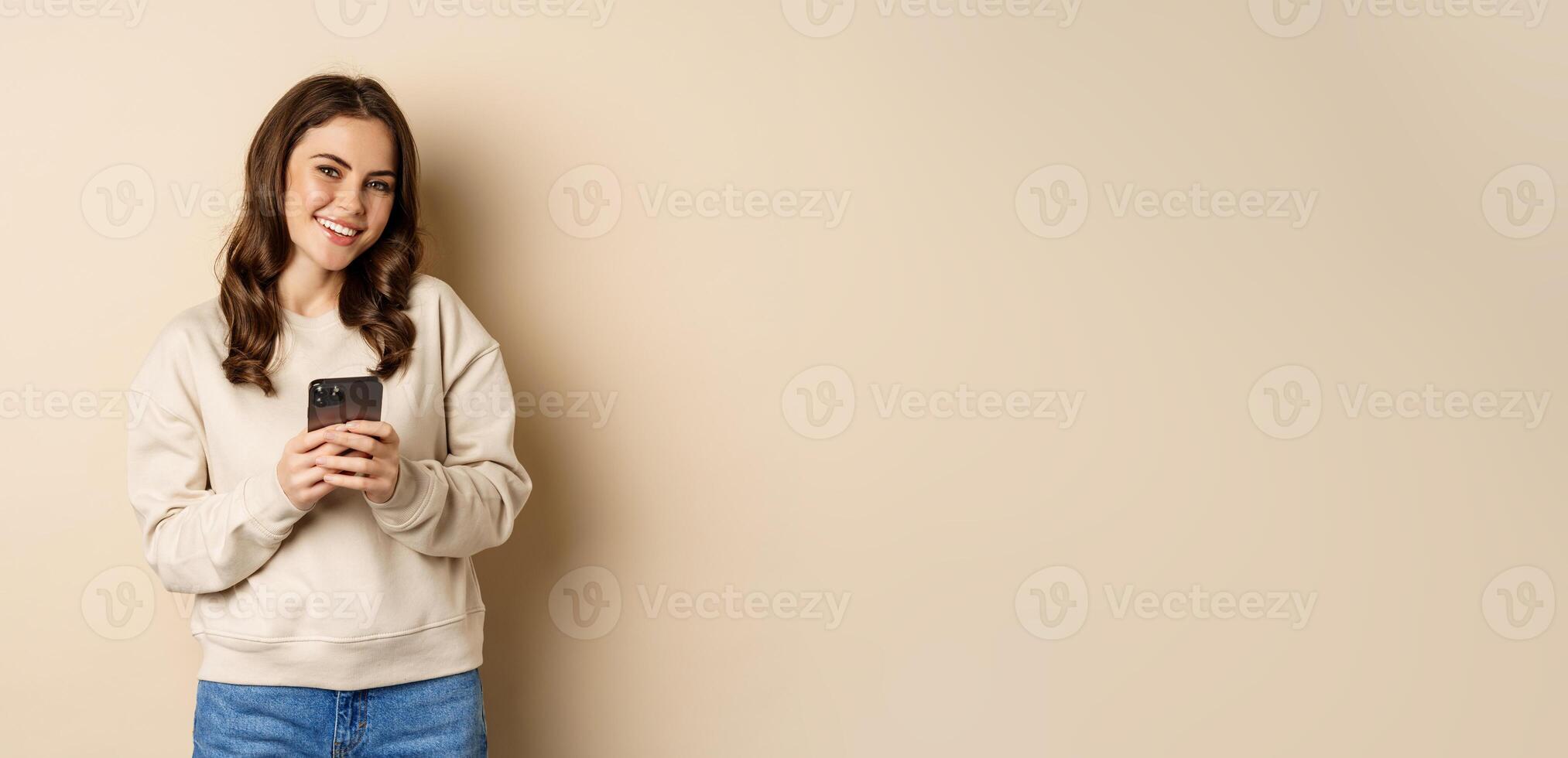 People and cellular technology. Beautiful stylish woman using mobile phone, smartphone app, smiling and looking at screen, standing over beige background photo