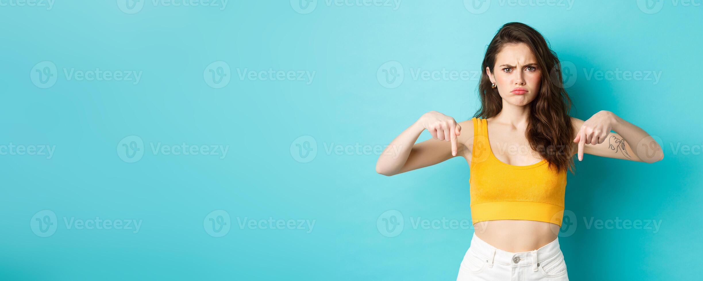 Confused frowning woman looking displeased, need explanation, pointing fingers down at something strange or unpleasant, standing over blue background photo