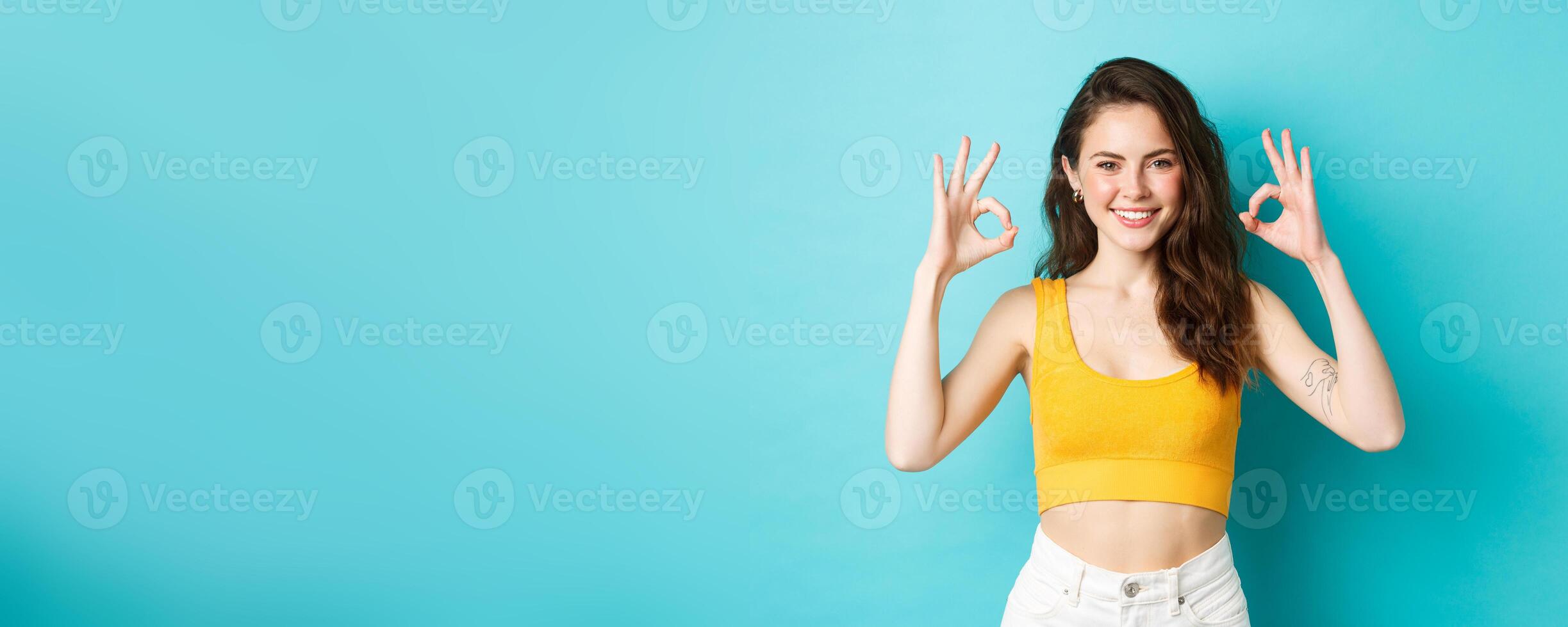 Beautiful and stylish confident woman showing okay signs, approve good choice, wearing summer clothes, standing over blue background photo