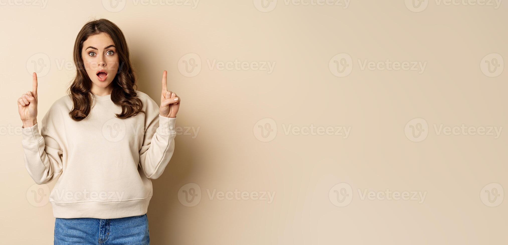Surprised beautiful girl showing advertisement, pointing fingers on top, store sale or website link up, standing over beige background photo