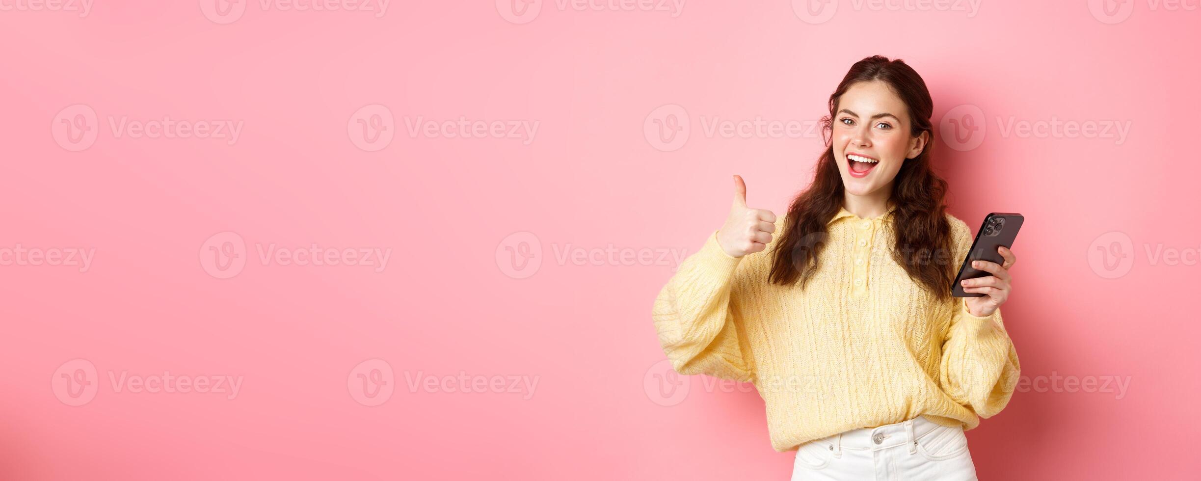 Portrait of attractive glamour girl showing thumbs up after using app on smartphone, online shopping application, standing against pink background, smiling satisfied photo