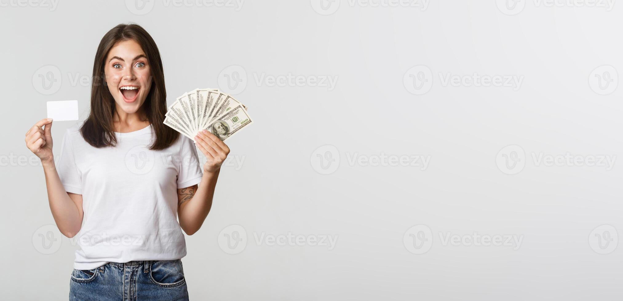 Excited happy brunette girl showing credit card and money, smiling over white background photo