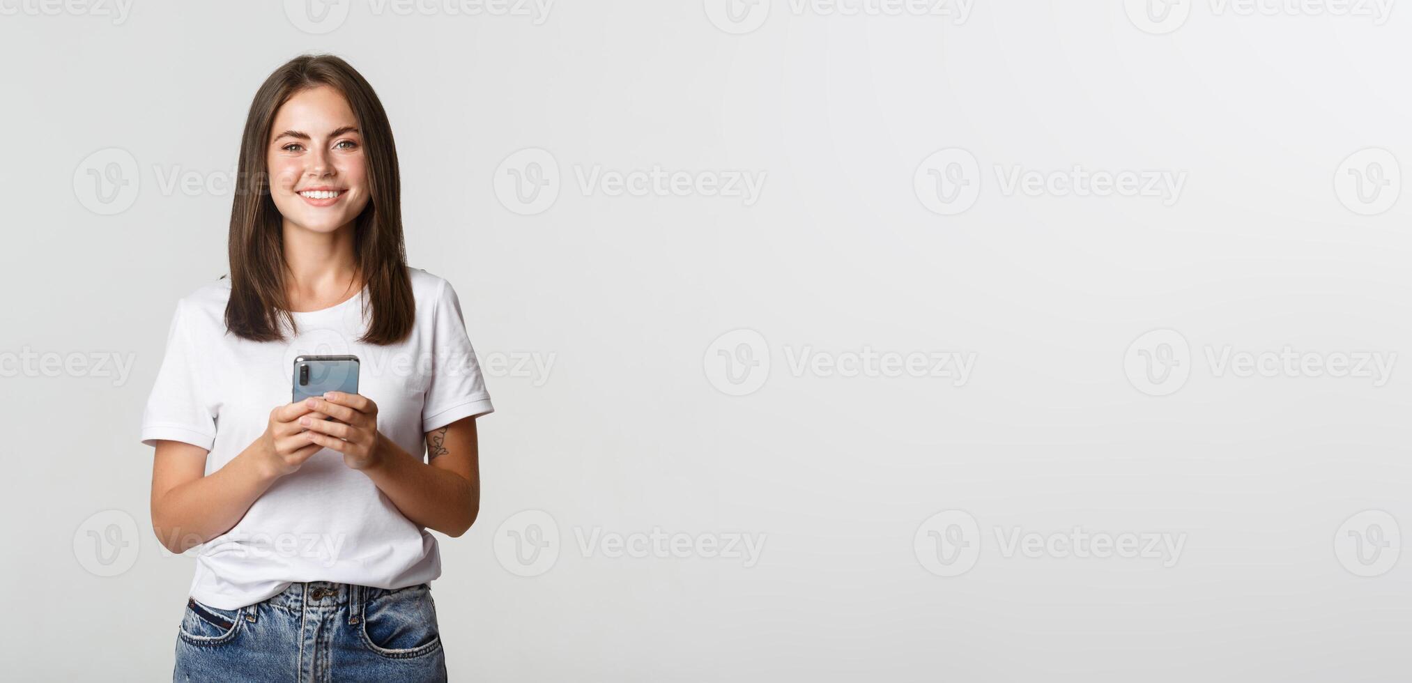 Attractive smiling brunette girl texting message, using mobile phone and looking at camera photo
