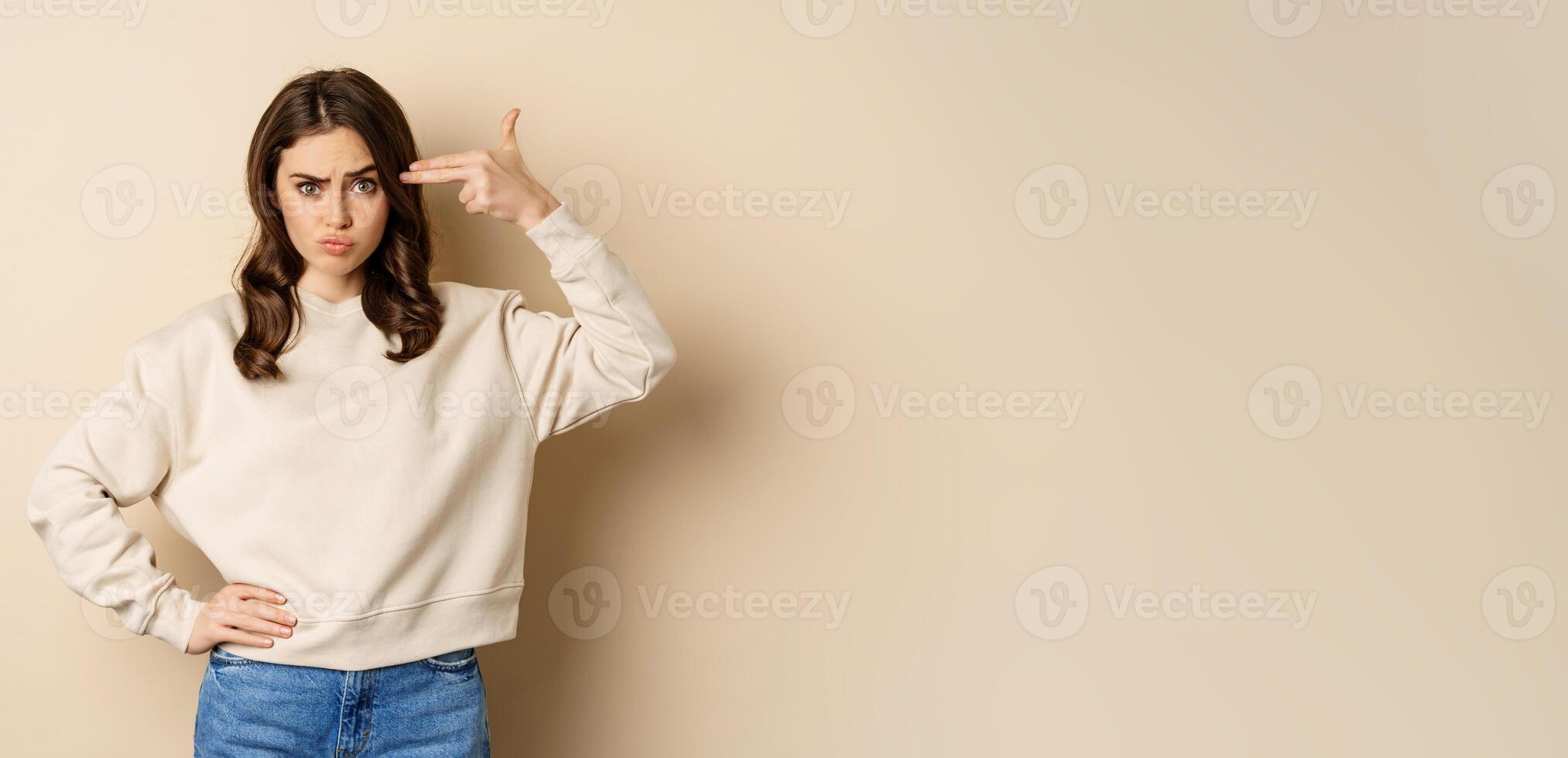 Woman showing finger pistol gesture over her head, being bothered and annoyed, standing over beige background photo