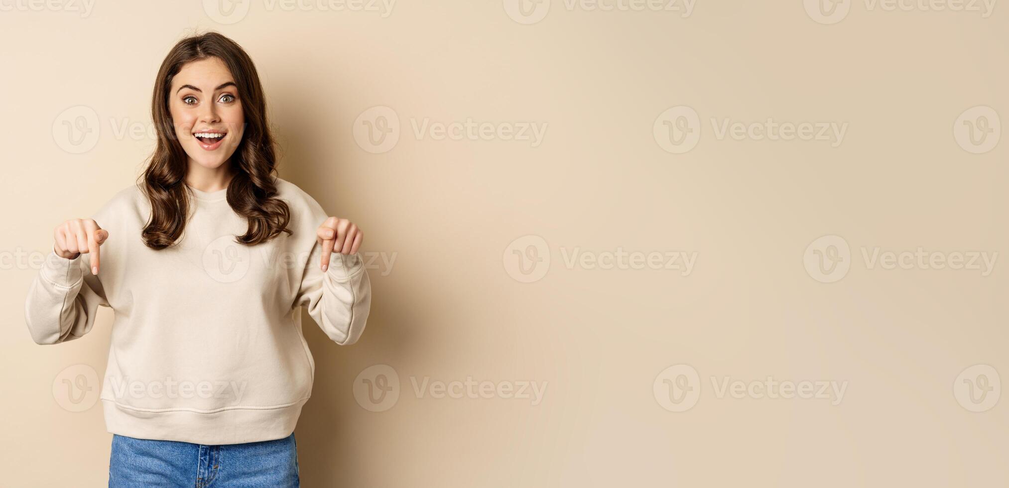 Enthusiastic attractive girl pointing fingers down, showing advertisement, link or promo below, standing over beige background photo