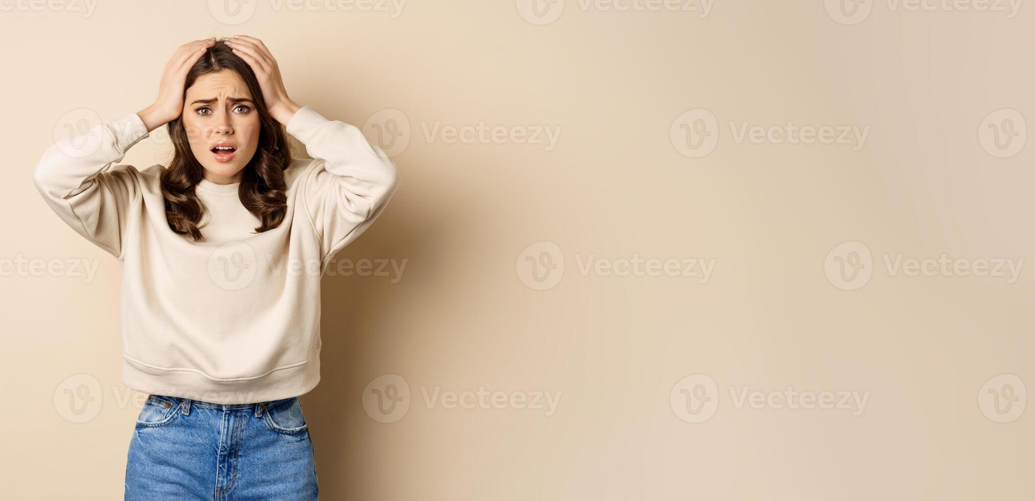 Frustrated and troubled woman facing disaster, holding hands on head in panic, looking anxious, standing over beige background photo