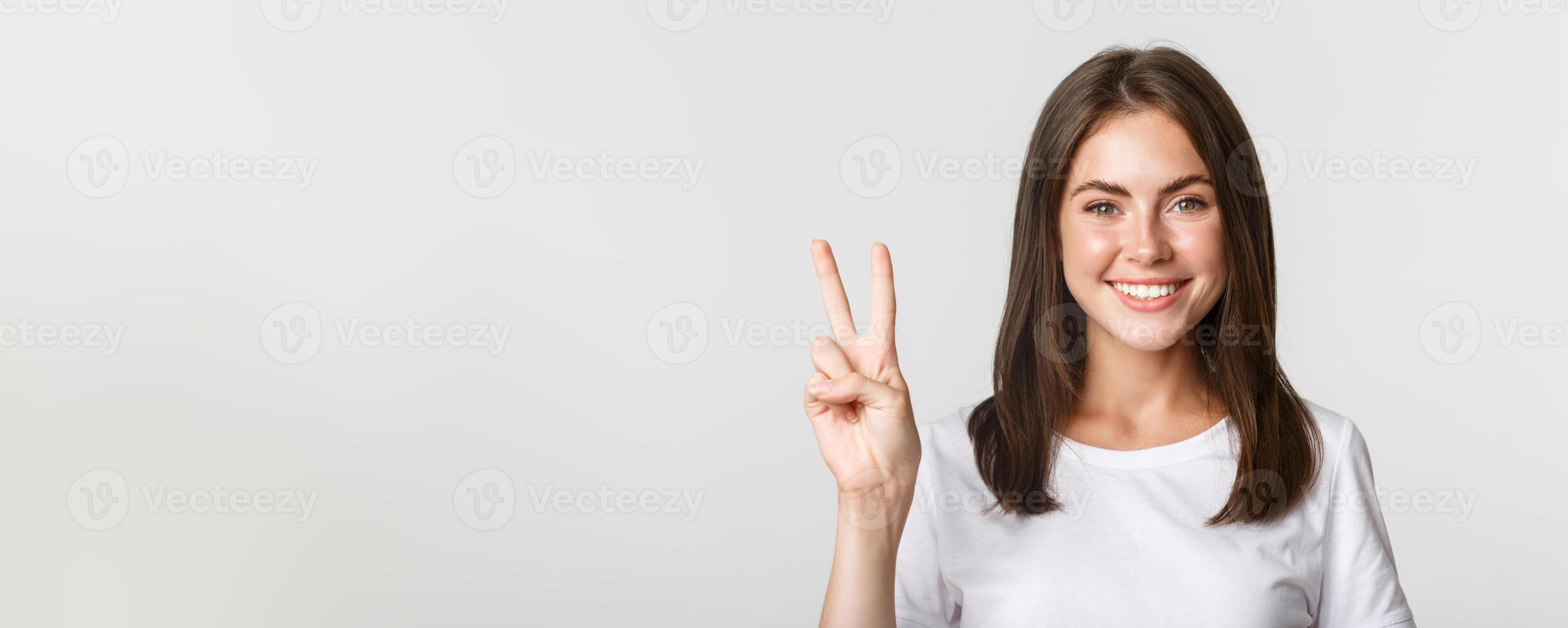 Portrait of beautiful and happy brunette girl smiling and showing peace gesture, standing white background photo