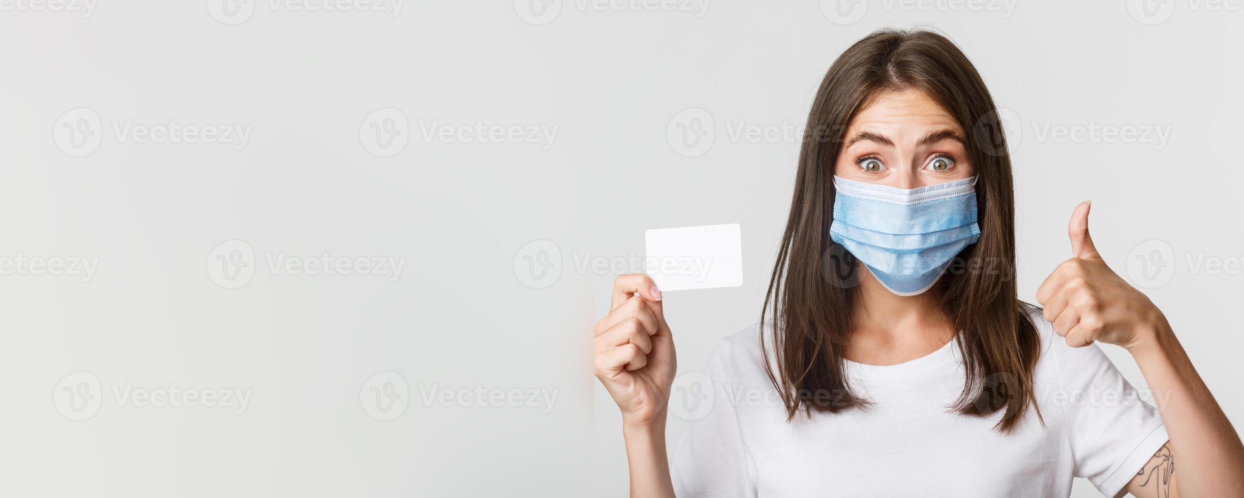 Covid-19, health and social distancing concept. Close-up of pretty brunette girl in medical mask, showing credit card and thumbs-up in approval photo