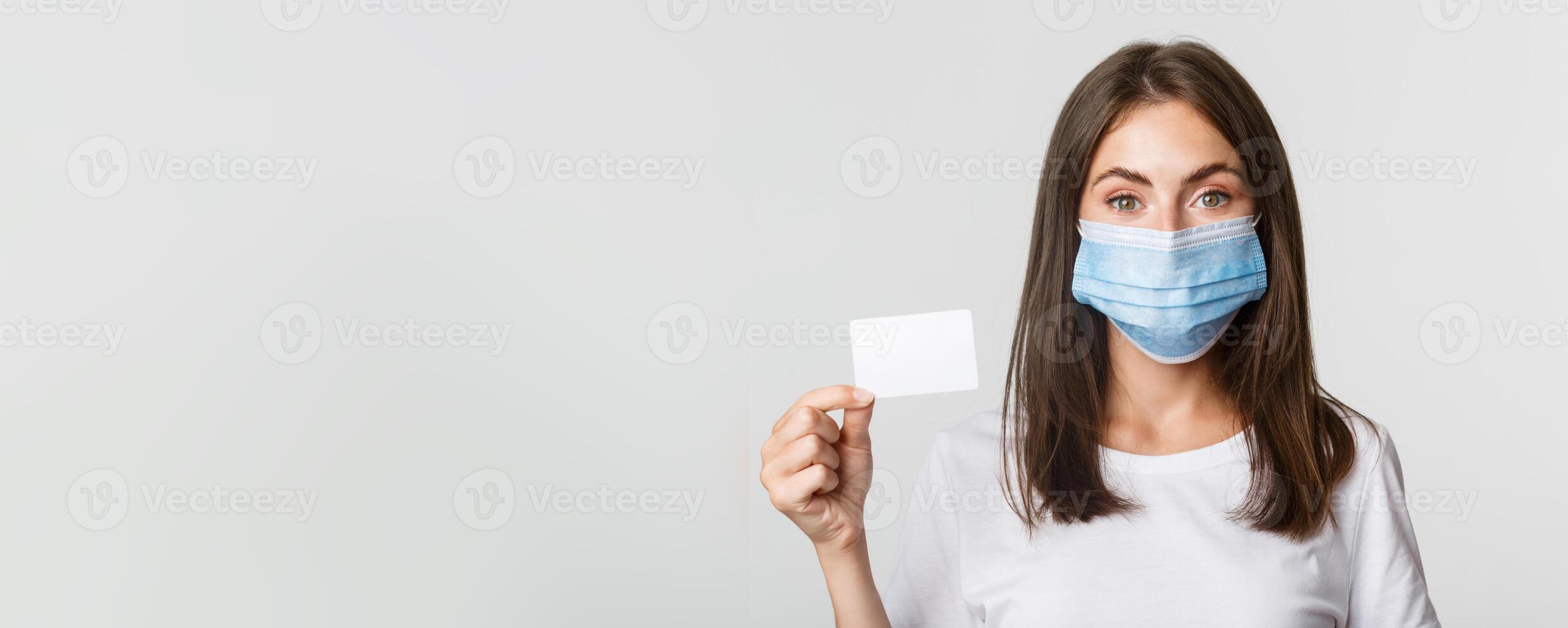 Covid-19, health and social distancing concept. Close-up of pretty brunette girl in medical mask, showing credit card photo