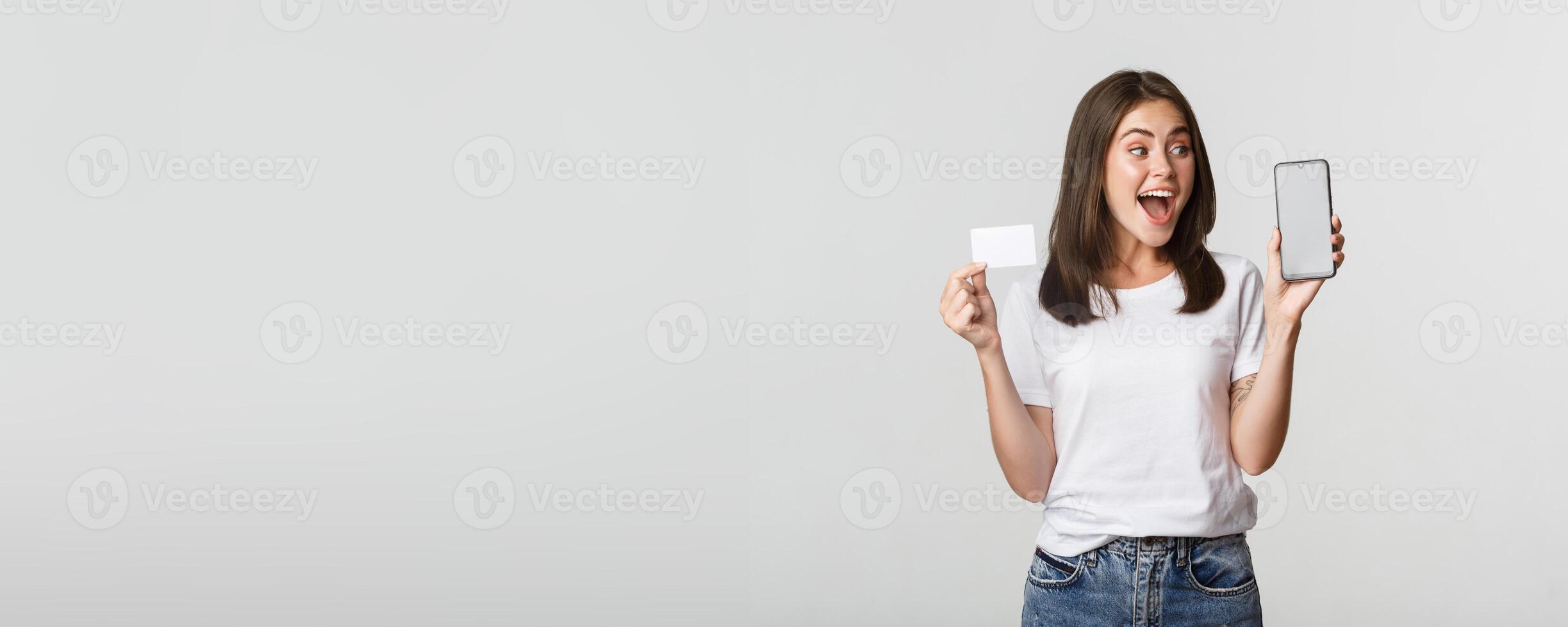 Excited and surprised cute girl showing credit card and mobile phone banking app on screen photo