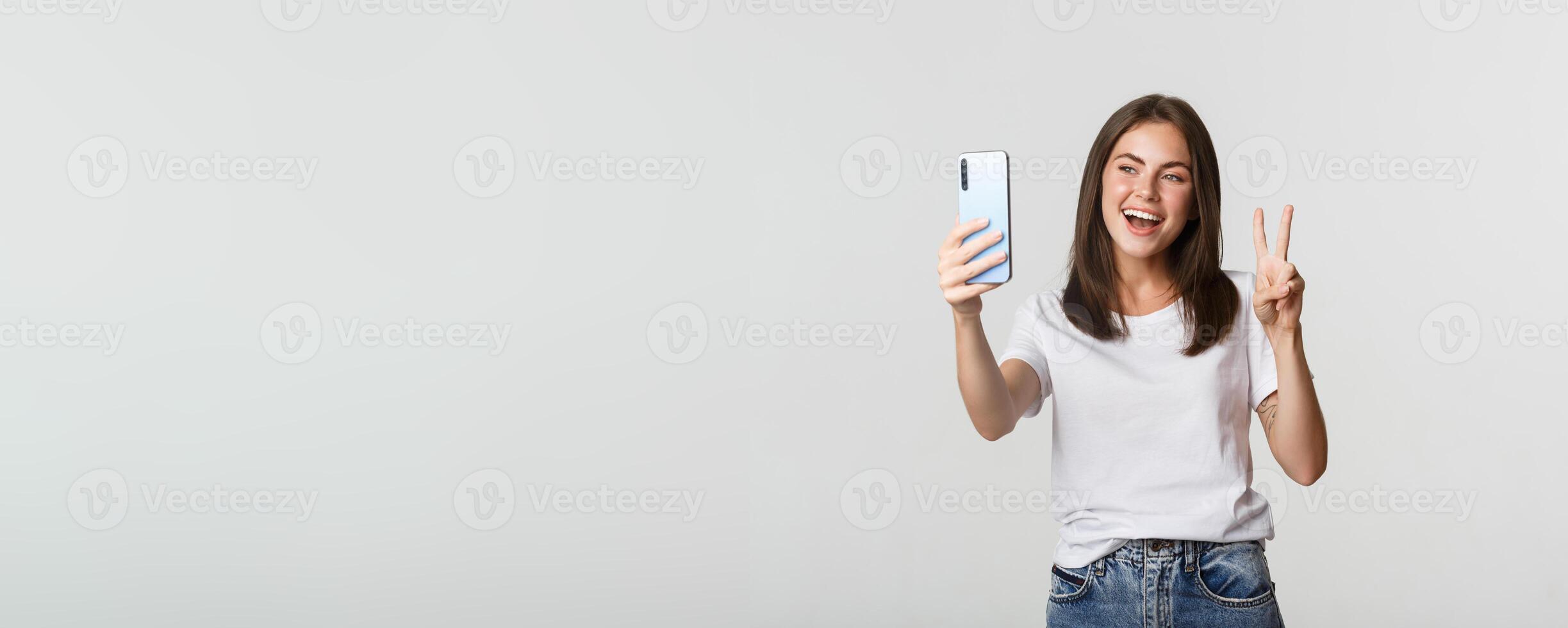 Attractive brunette girl looking happy and taking selfie on smartphone, showing peace gesture photo