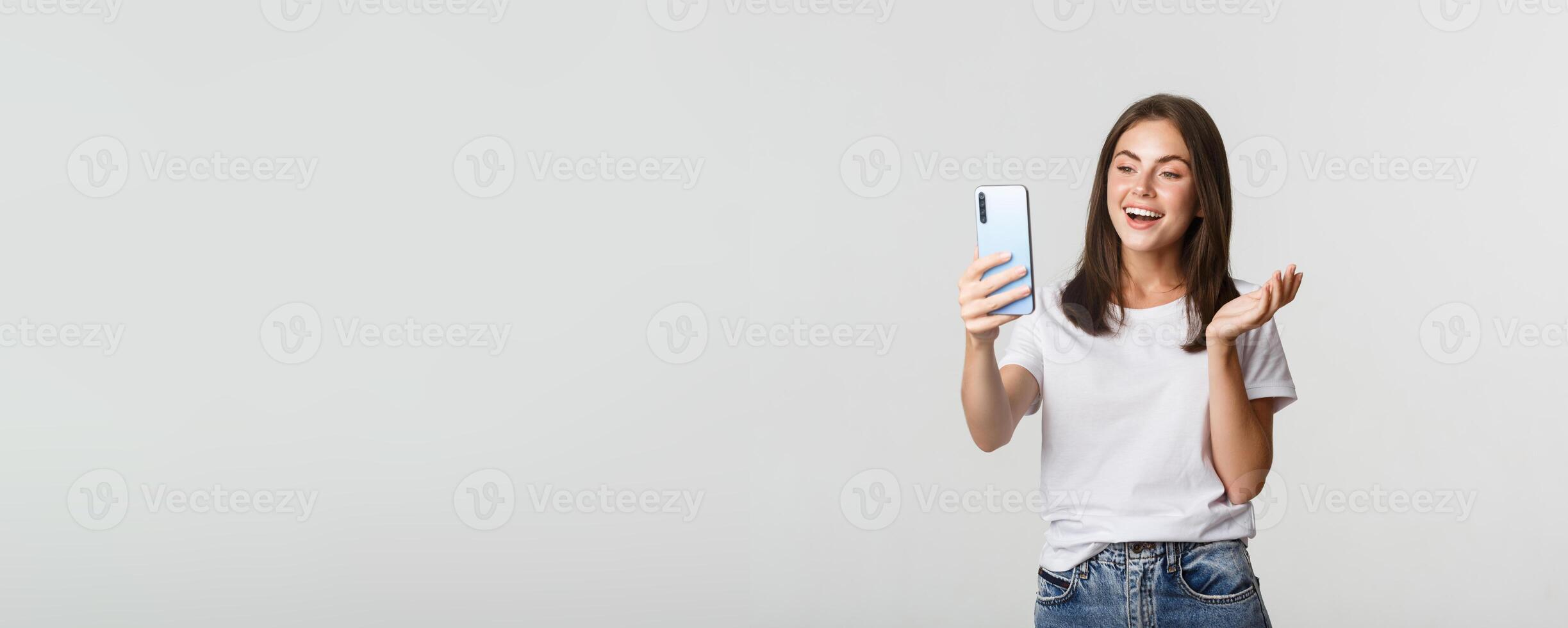 Friendly attractive girl calling friend, smiling and having conversation, holding smartphone, white background photo