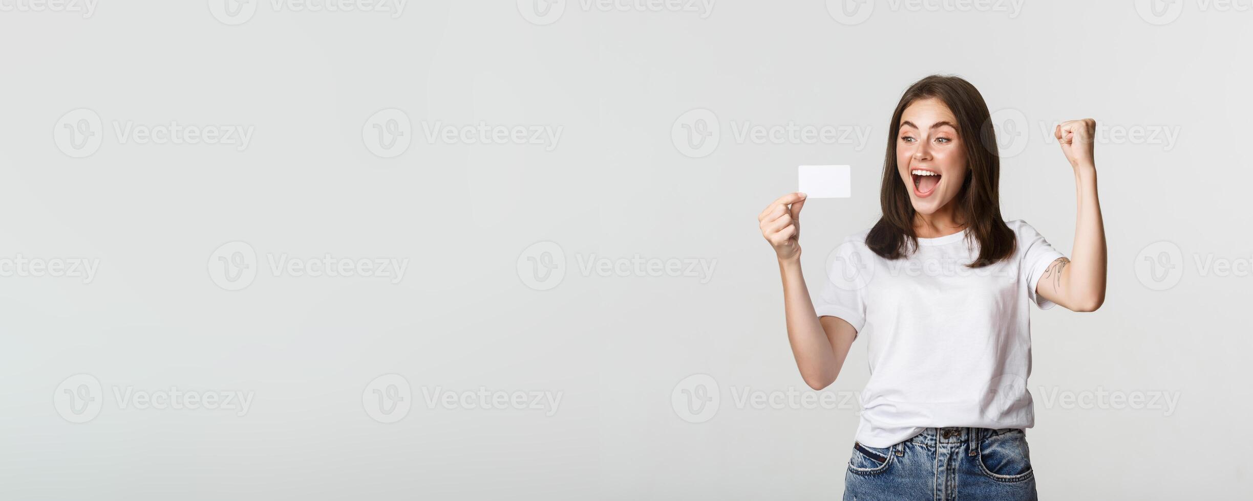Joyful good-looking girl rejoicing and looking at credit card, fist pump while triumphing, white background photo