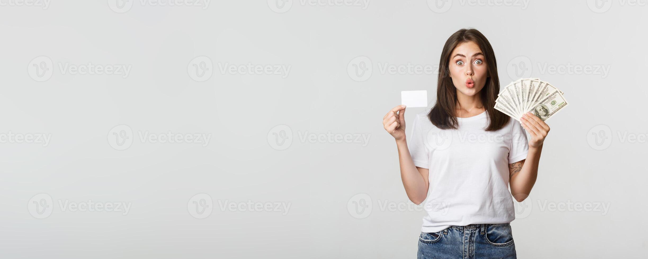 Amused brunette girl looking excited, holding money and credit card, standing white background photo