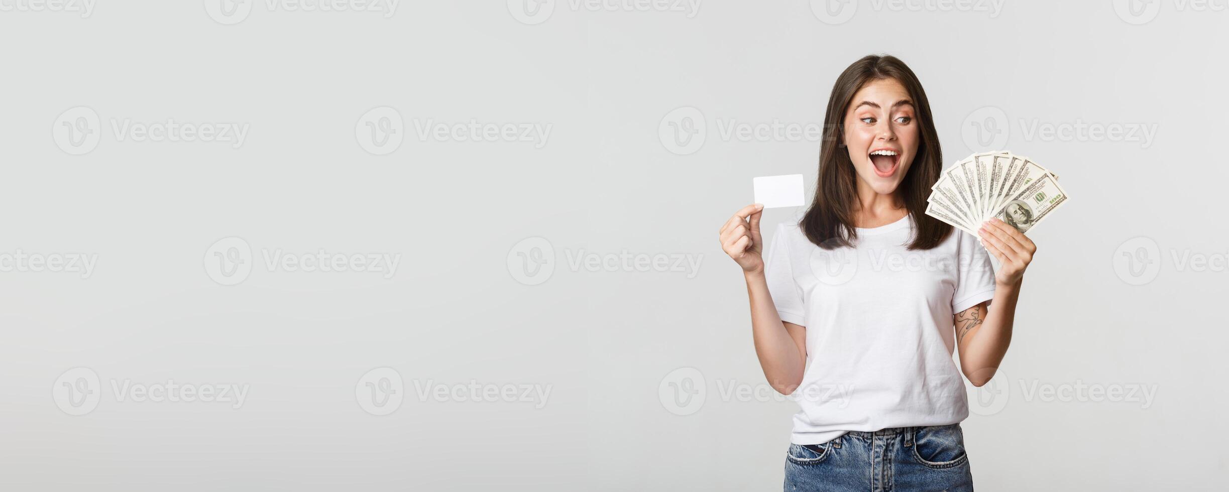 Amused beautiful girl looking at money and holding credit card, standing white background photo