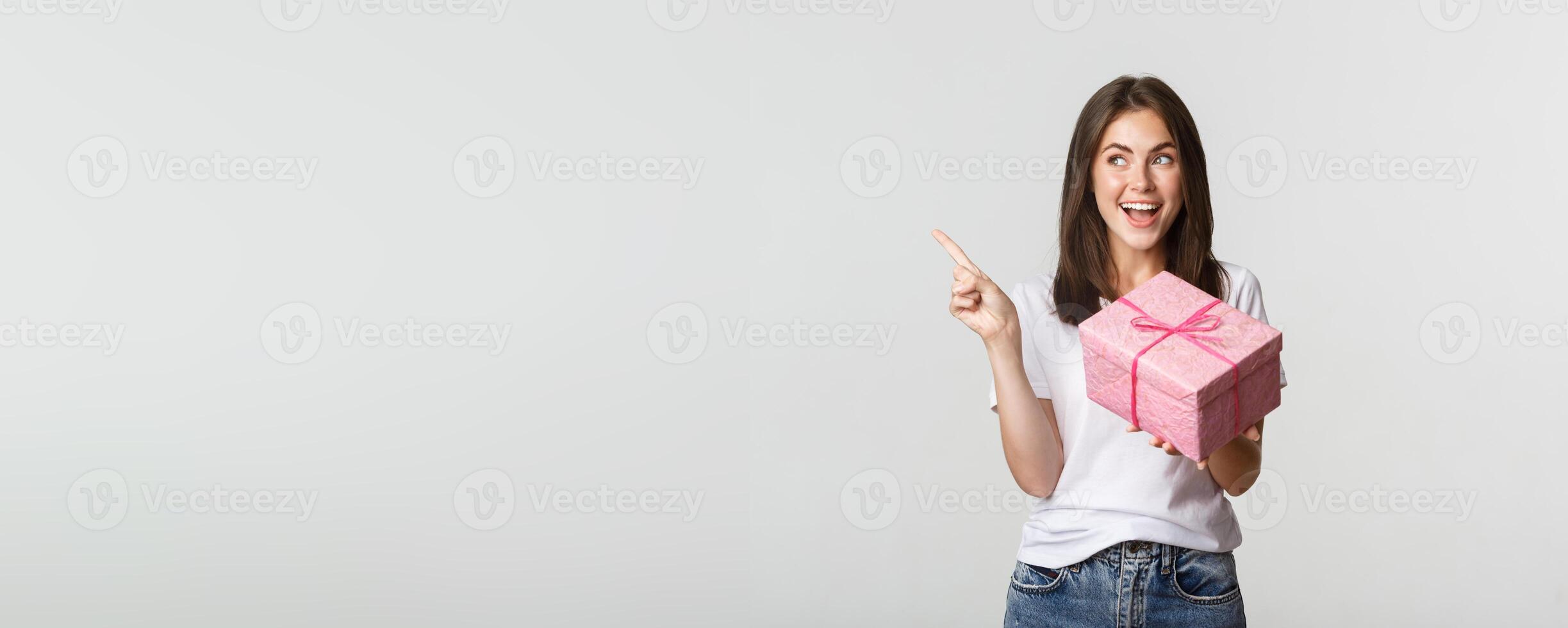 Happy smiling brunette girl holding birthday gift and pointing finger left at logo photo