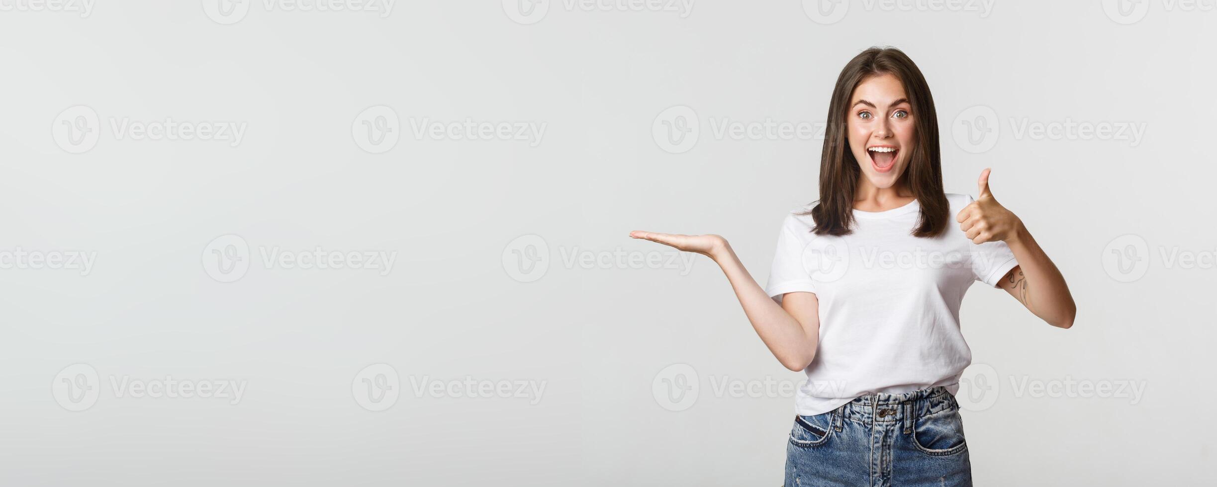 Satisfied attractive girl holding something on hand and showing thumbs-up in approval photo