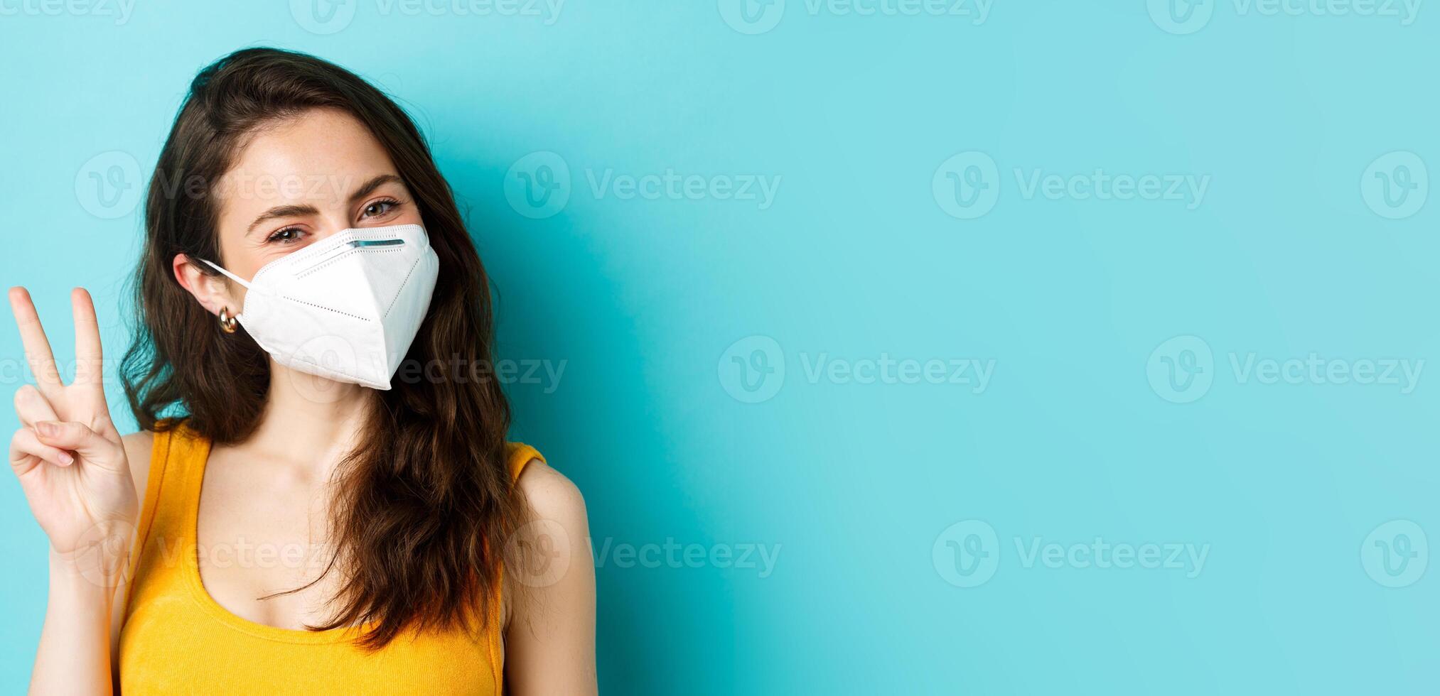 Covid-19, coronavirus and social distancing. Young positive woman in respirator showing v-sign, standing against blue background photo