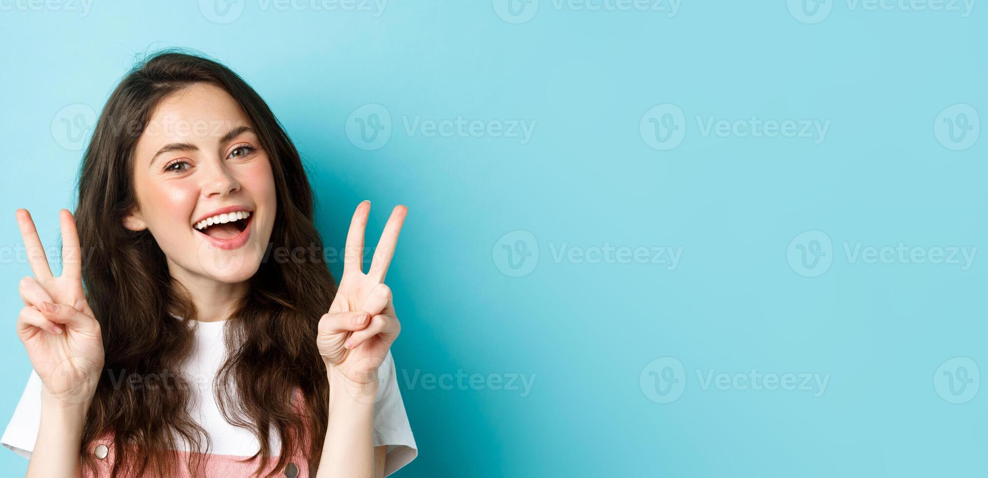 Young positive modern girl sending good vibes only, showing v-sign and smiling happy, standing against blue background photo