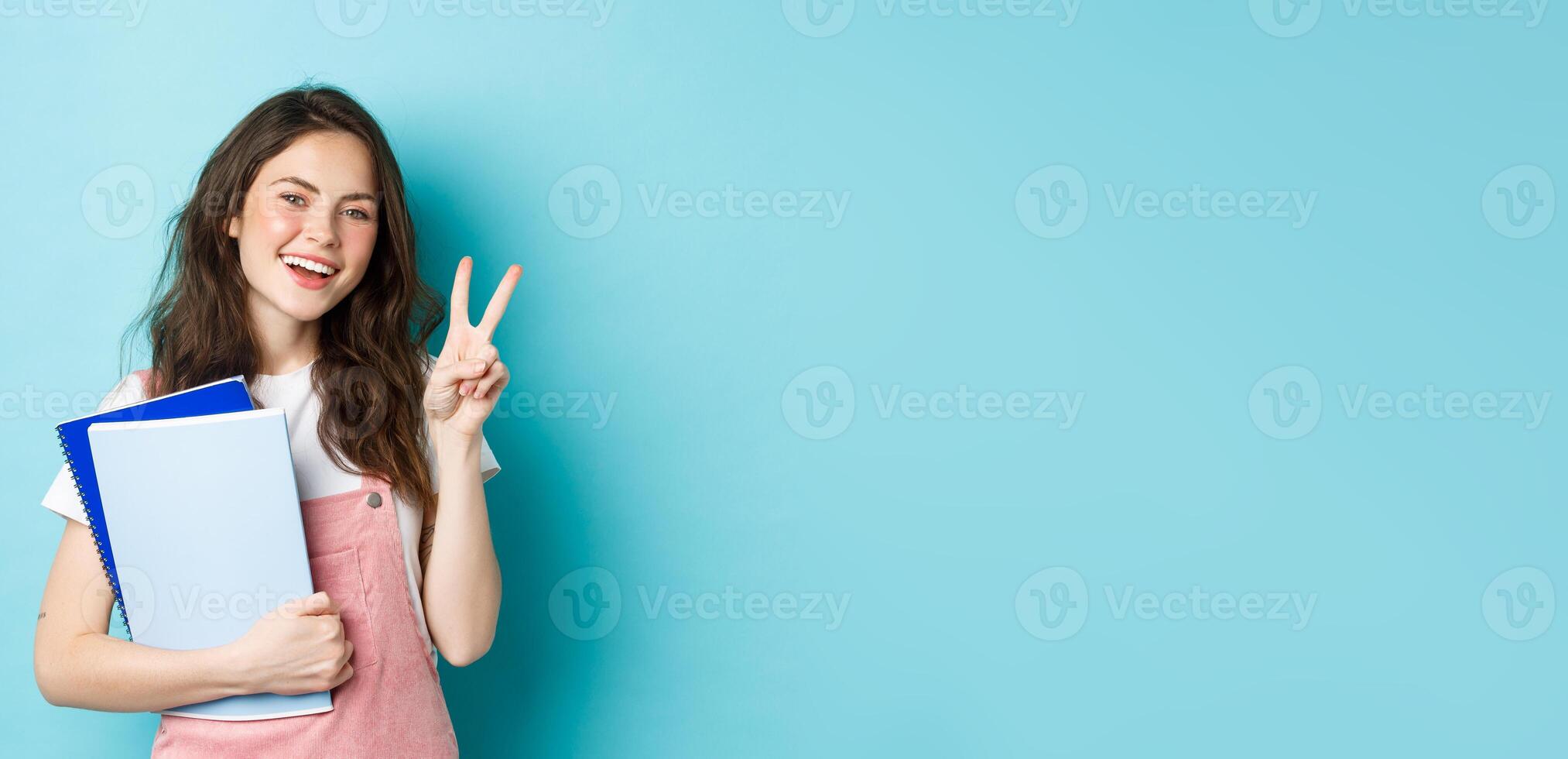 Beautiful female student showing v-sign and smiling happy, holding notebooks with study material, attending courses, standing over blue background photo