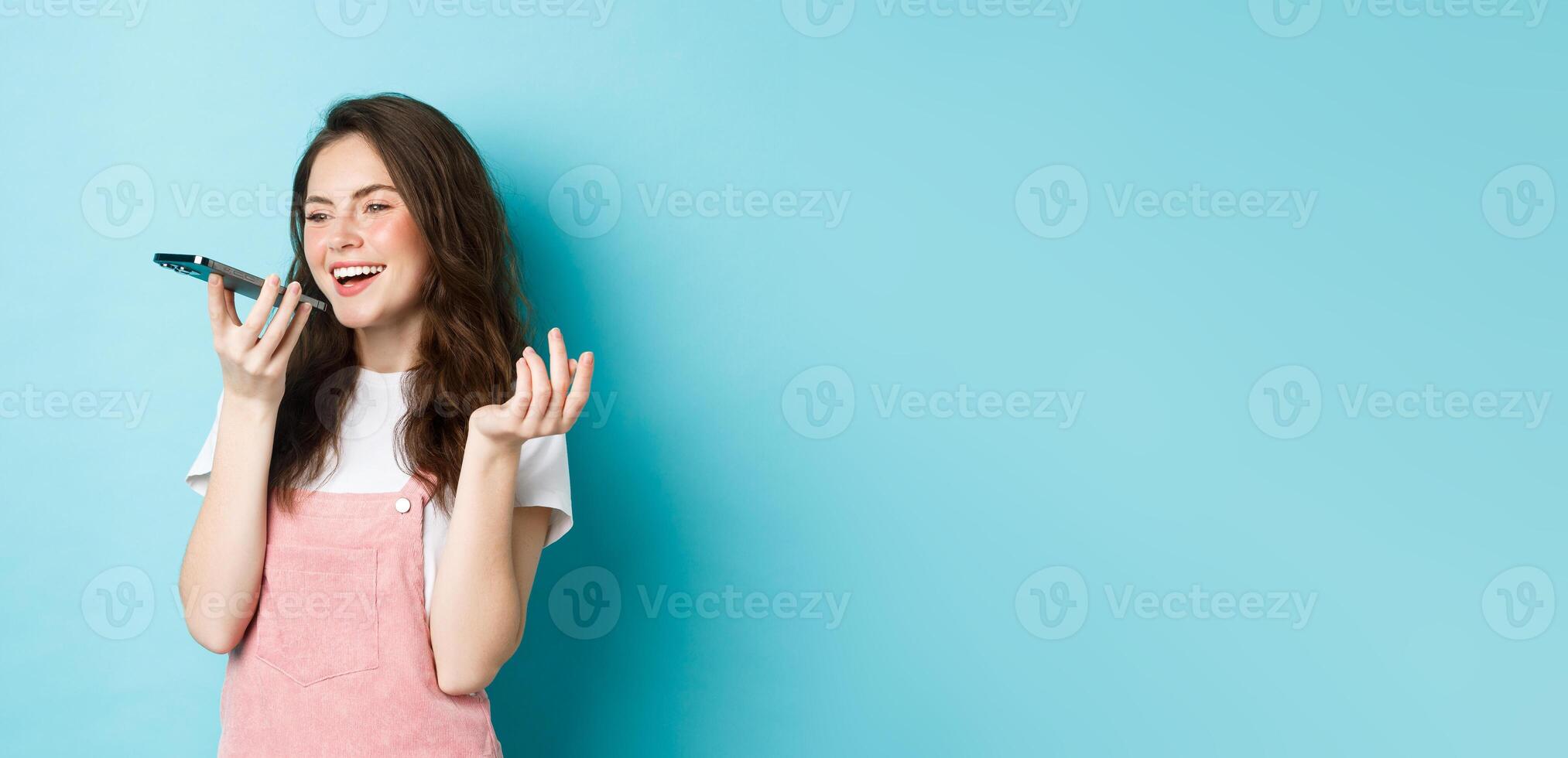 Portrait of smiling woman holding phone near lips and talking, using app translator on smartphone or recording voice message, speaking with speakerphone, standing over blue background photo