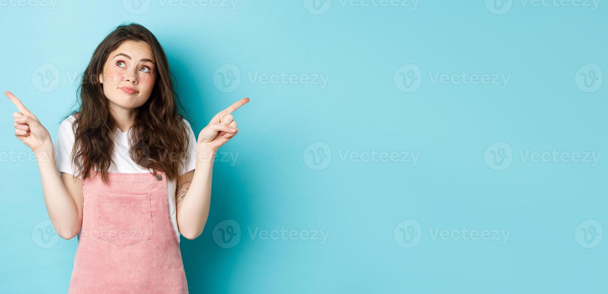 Indecisive beautiful girl thinking, making her choice, pointing sideways at left and right sides and shrugging confused, dont know what to pick or buy, standing over blue background photo