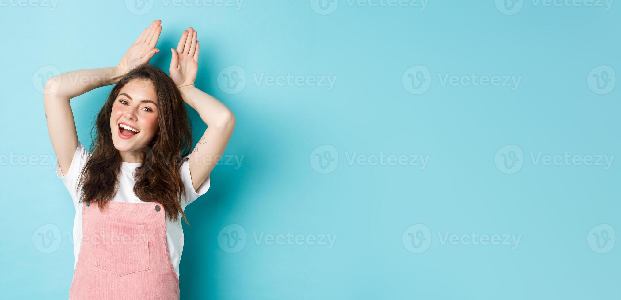 Beautiful girl mimic Easter bunny, showing rabbit ears with hands on head and smiling lovely, dancing happy, standing over blue background photo