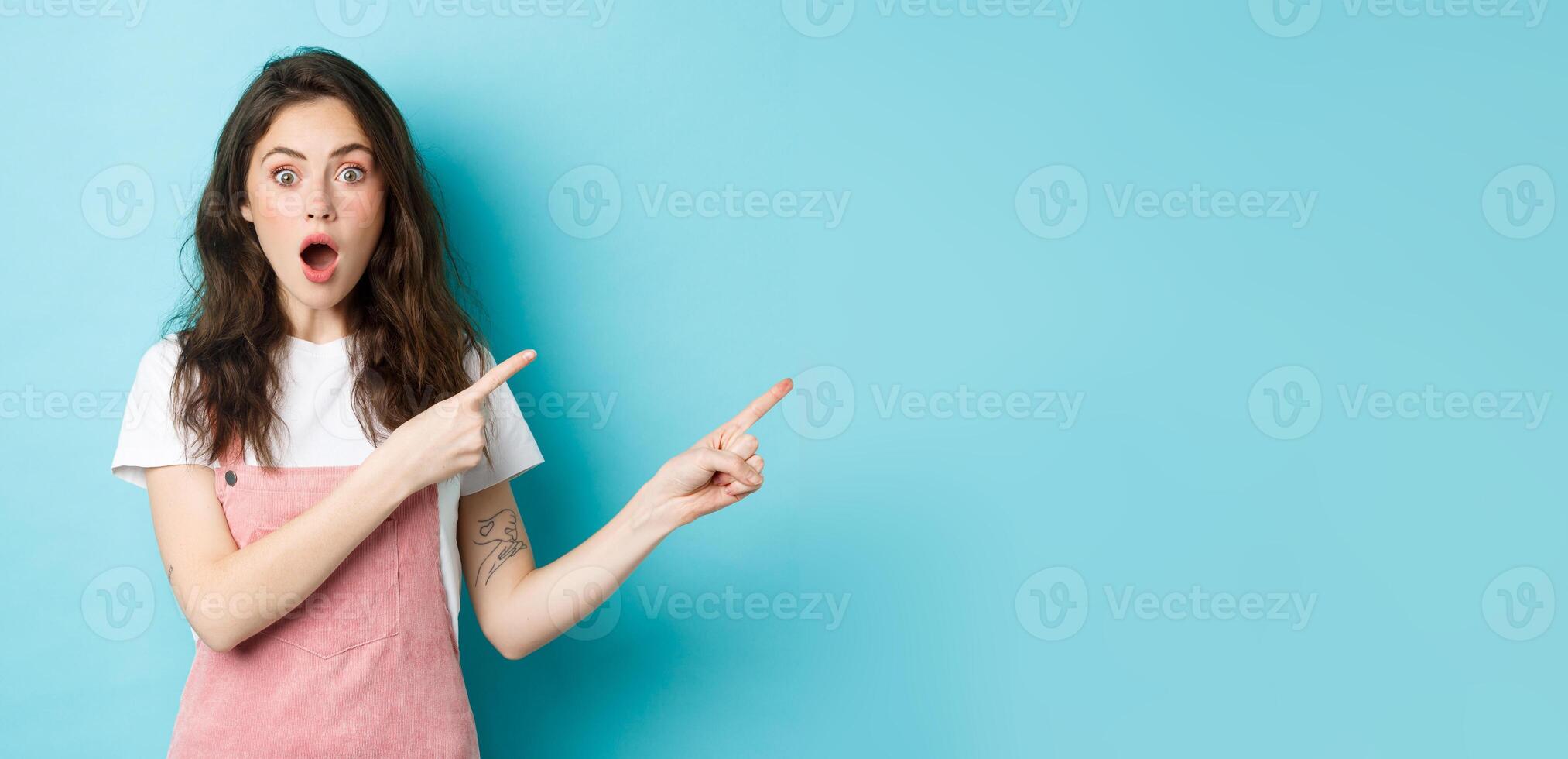 Excited and surprised young woman open mouth, say wow and pointing fingers right at logo, staring impressed at camera while talking about new product or news, blue background photo