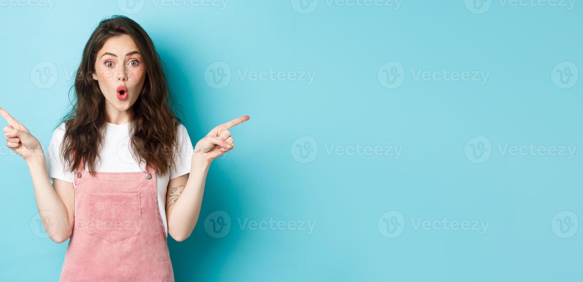 Wow check it out. Excited brunette girl with glamour summer make up, pointing fingers sideways at left and right sides, gasping amazed, standing over blue background photo