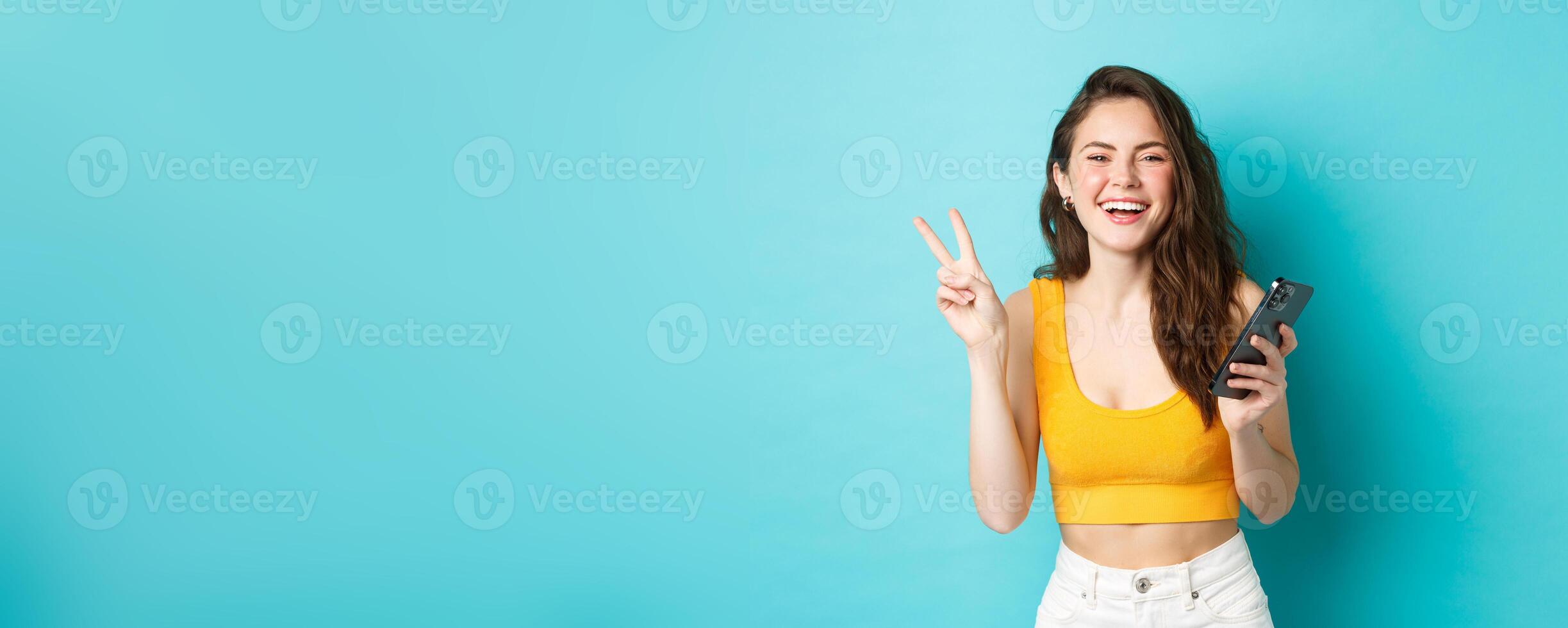 Technology and lifestyle concept. Happy attractive woman laughing, showing v-sign and using smartphone, chatting on mobile phone, standing over blue background photo