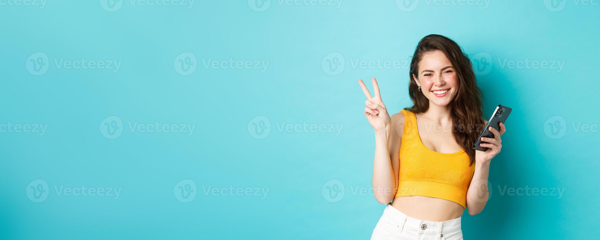 Technology and lifestyle concept. Beautiful cheerful girl sending positive vibes, smiling and showing peace sign, using cellphone, standing against blue background photo
