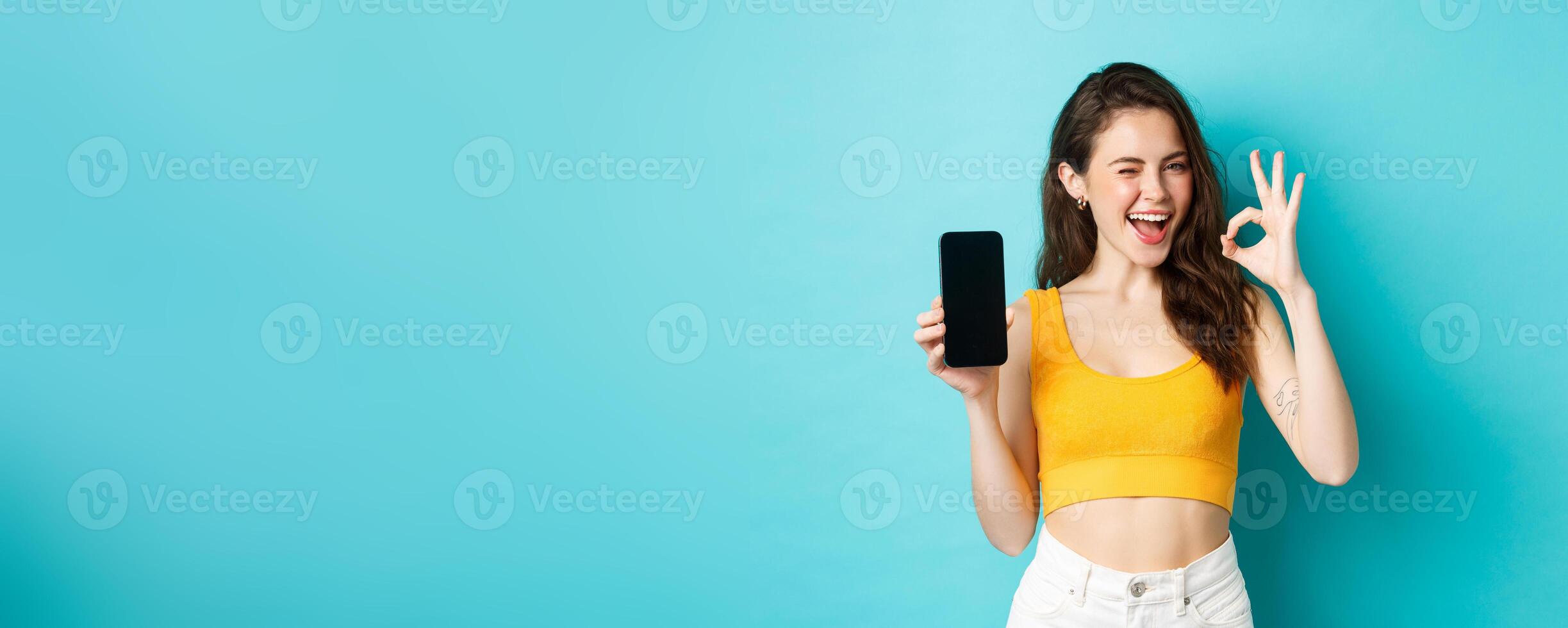 Attractive modern woman in summer clothes, winking assuringly, showing okay sign with blank smartphone screen, showing your logo on display, blue background photo