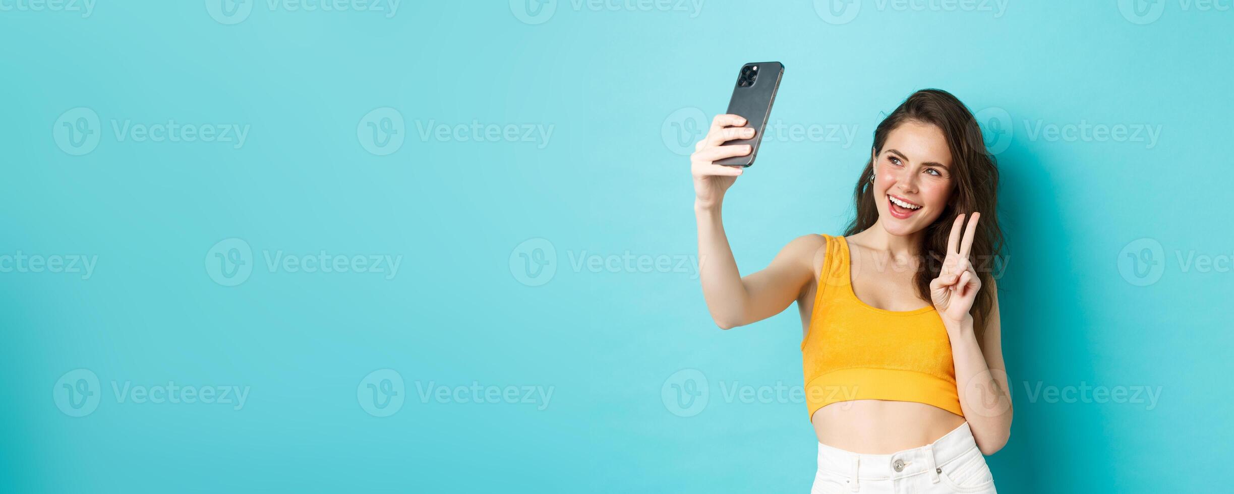 Technology and lifestyle concept. Stylish young woman taking selfie in her summer clothes, showing v-sign at smartphone camera, standing over blue background photo