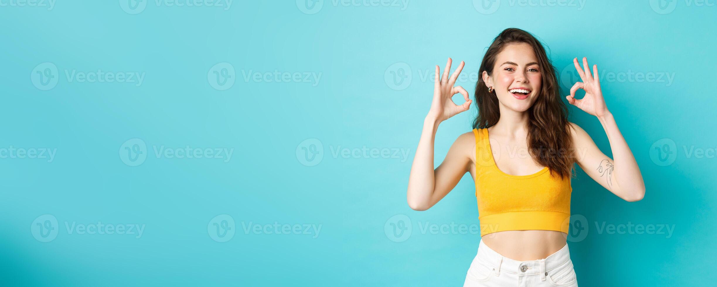 Young carefree woman say yes and nod in approval, showing okay gestures to praise good choice, show perfect body in summer outfit, ready for vacation, standing over blue background photo