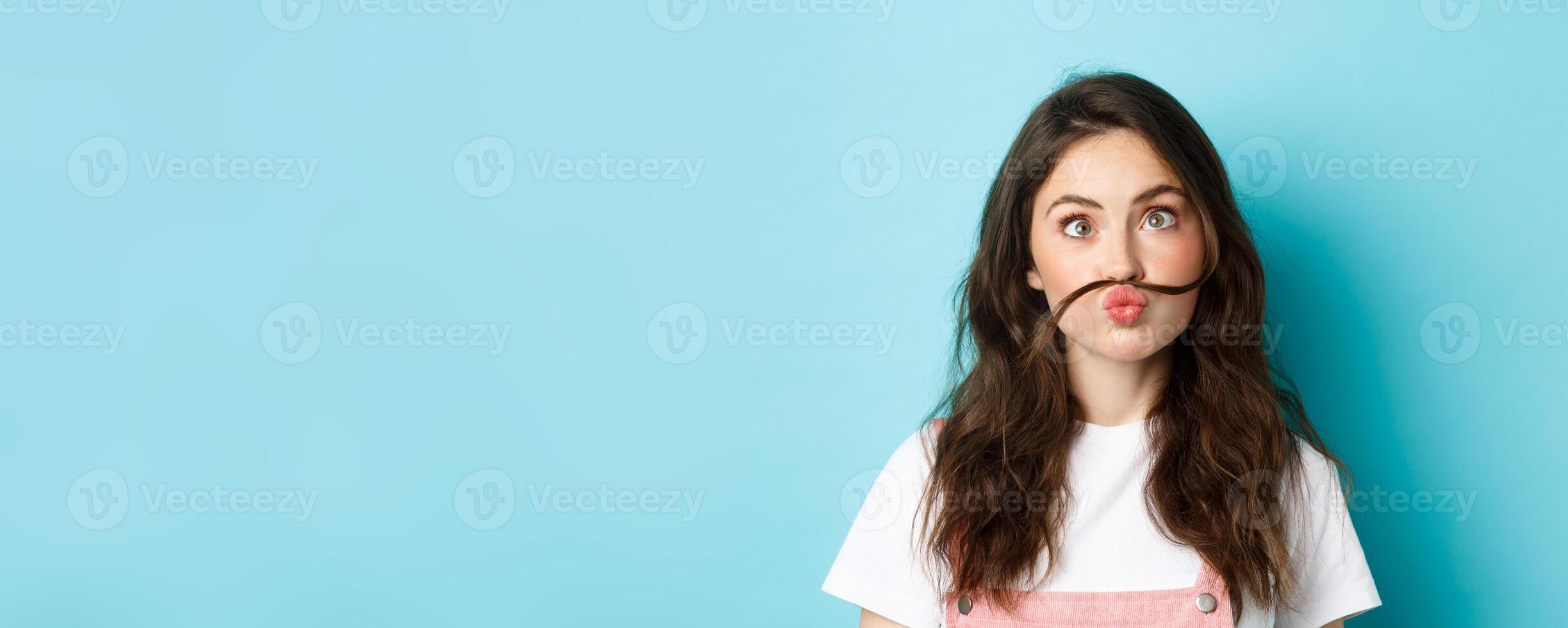 Close up portrait of funny silly girl playing, making squinting face and moustache with hair strand above lips, standing over blue background photo