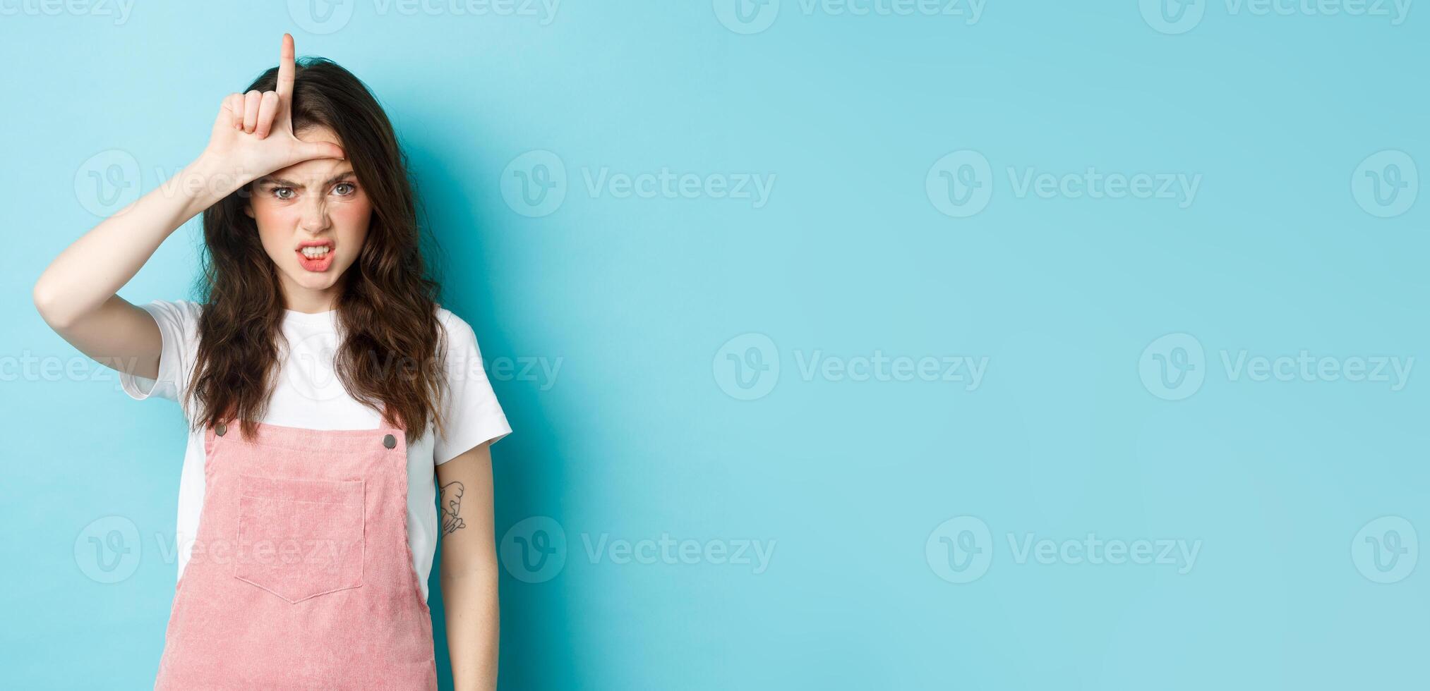Sassy young woman despise her rival, showing loser gesture on forehead, making letter L with hand and staring with dismay at camera, standing against blue background photo