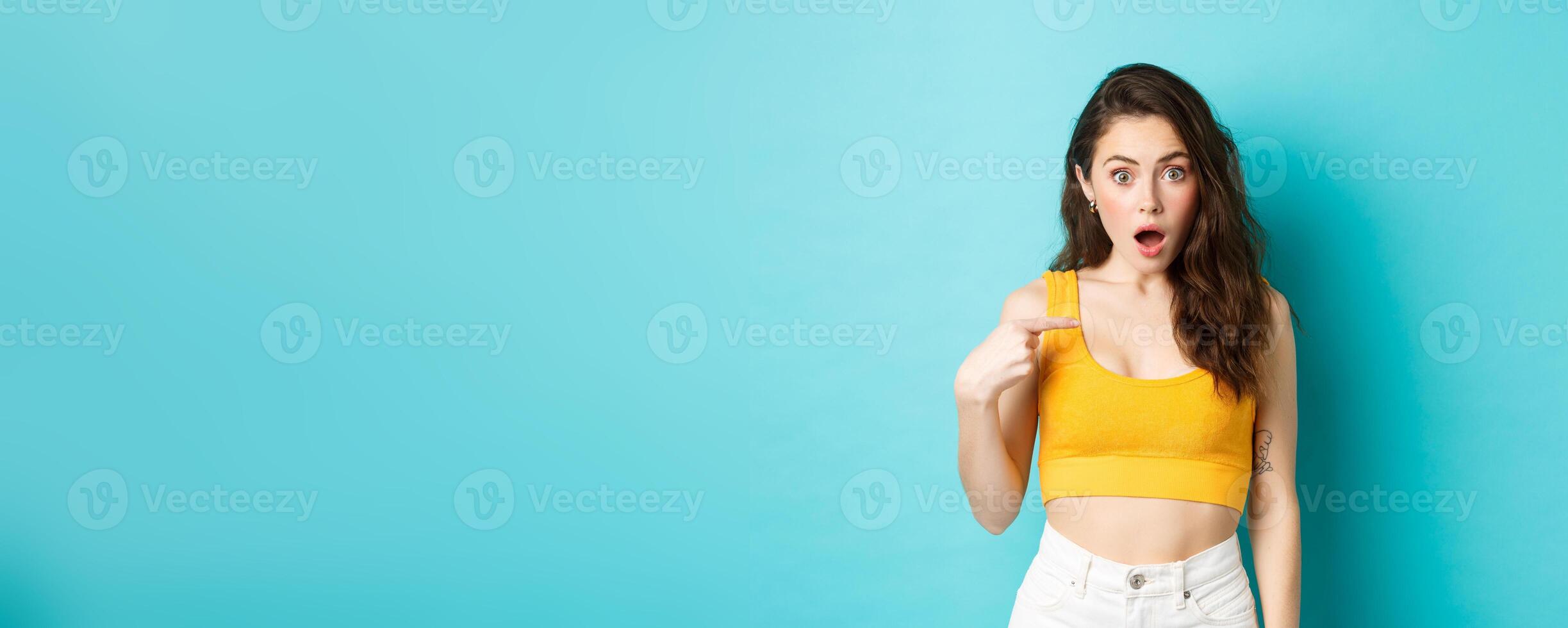 Portrait of young confused woman pointing at herself, looking with surprise and disbelief at camera, being accused or chosen unexpectedly, standing over blue background photo