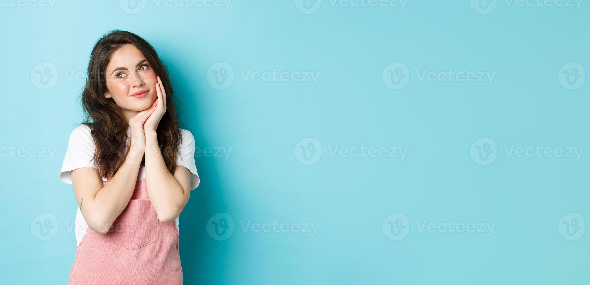 Portrait of beautiful girl dreaming, looking aside at logo banner and thinking, standing romantic against blue background, smiling and imaging something cute, blue background photo