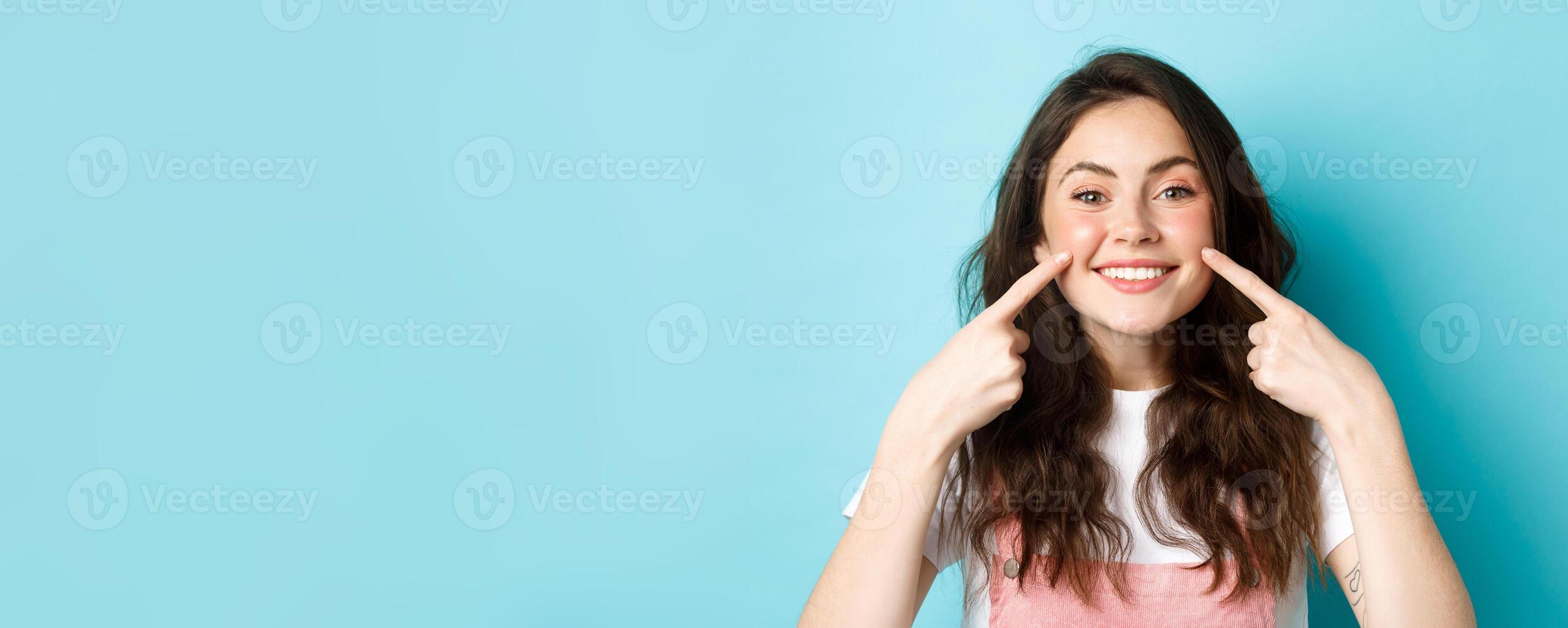 Portrait of happy young woman showing her clean glowing skin and blush on cheeks, pointing fingers at face, smiling satisfied at camera, blue background photo