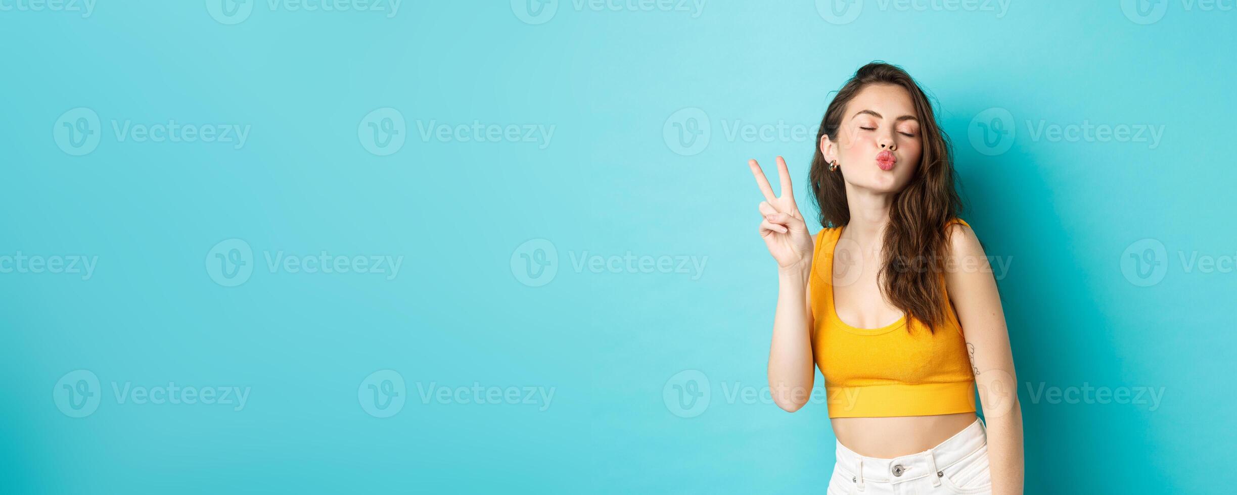 Glamour girl in summer clothes close her eyes and making kissing face with v-sign, enjoying vacation, having fun, standing relaxed against blue background photo