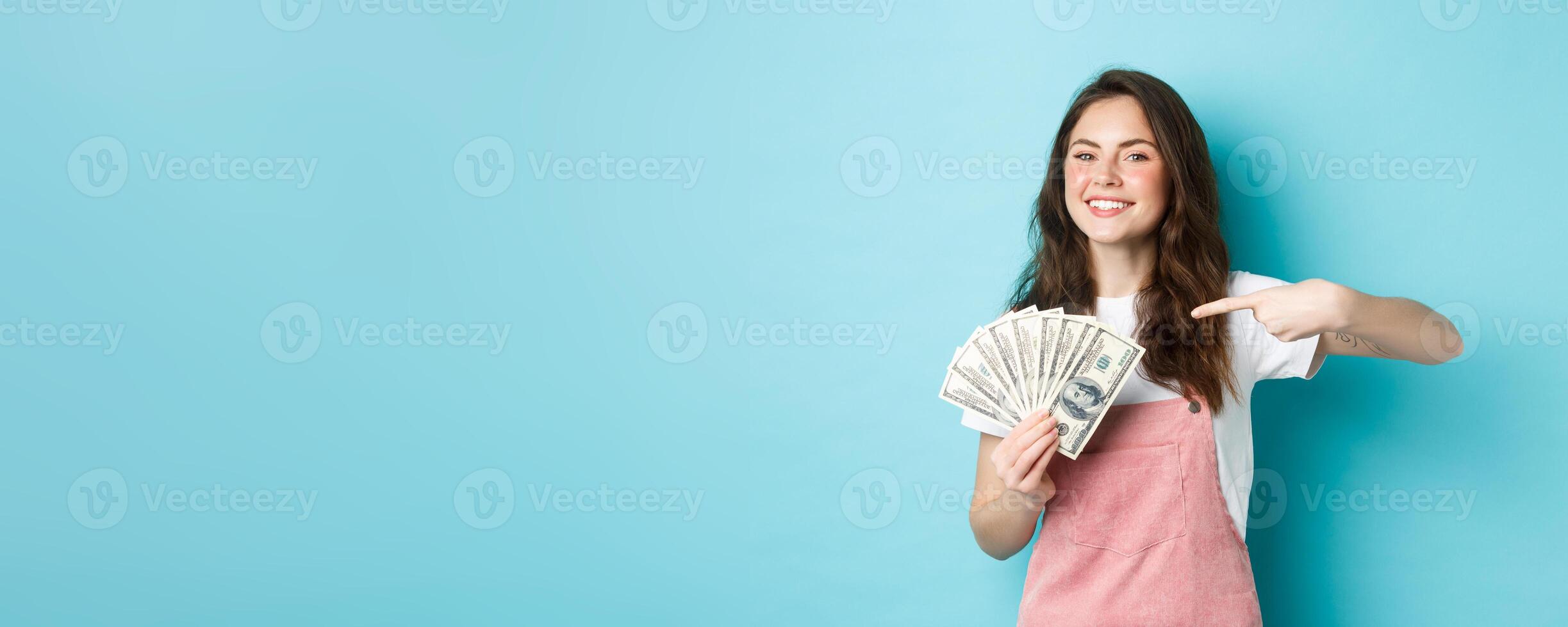 Portrait of cute girl smiling proud and satisfied, pointing finger at money dollar, showing dollar bills, standing over blue background photo