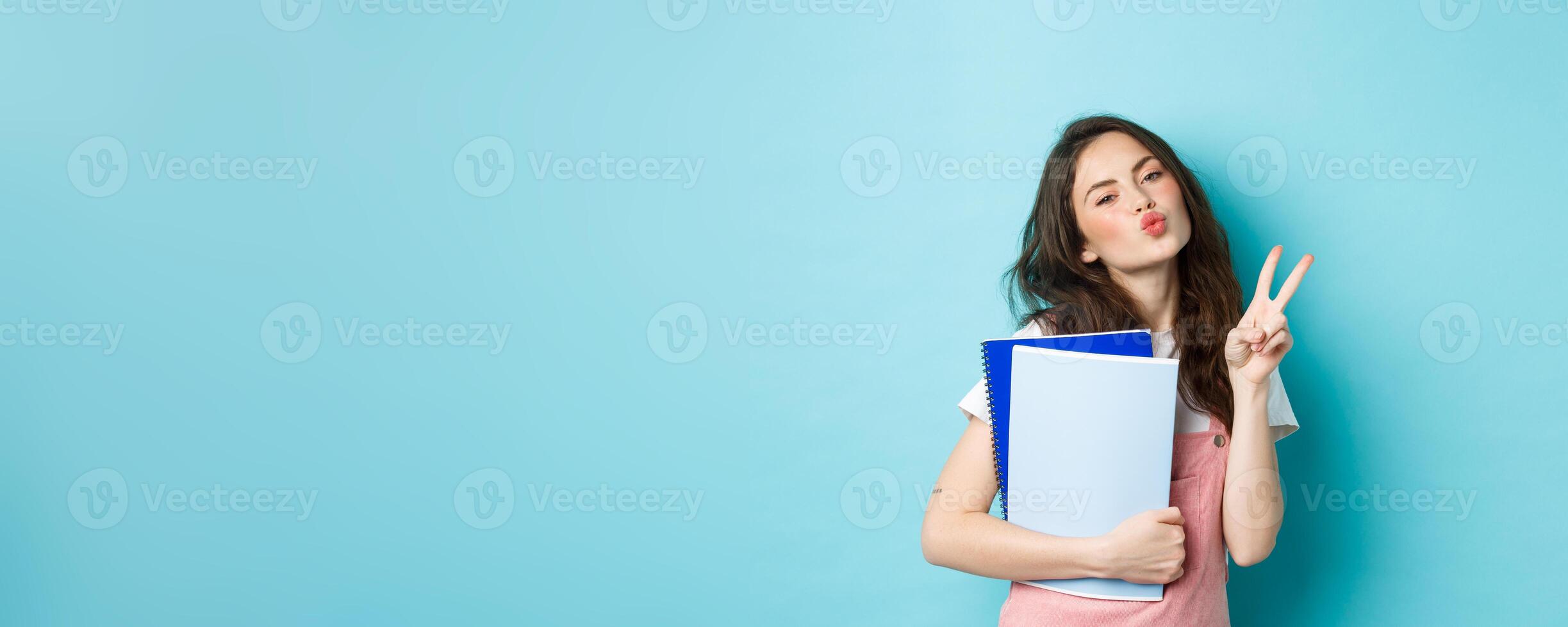 Portrait of glamour girl showing kissing face and v-sign, carry notebooks homework material, standing over blue background photo