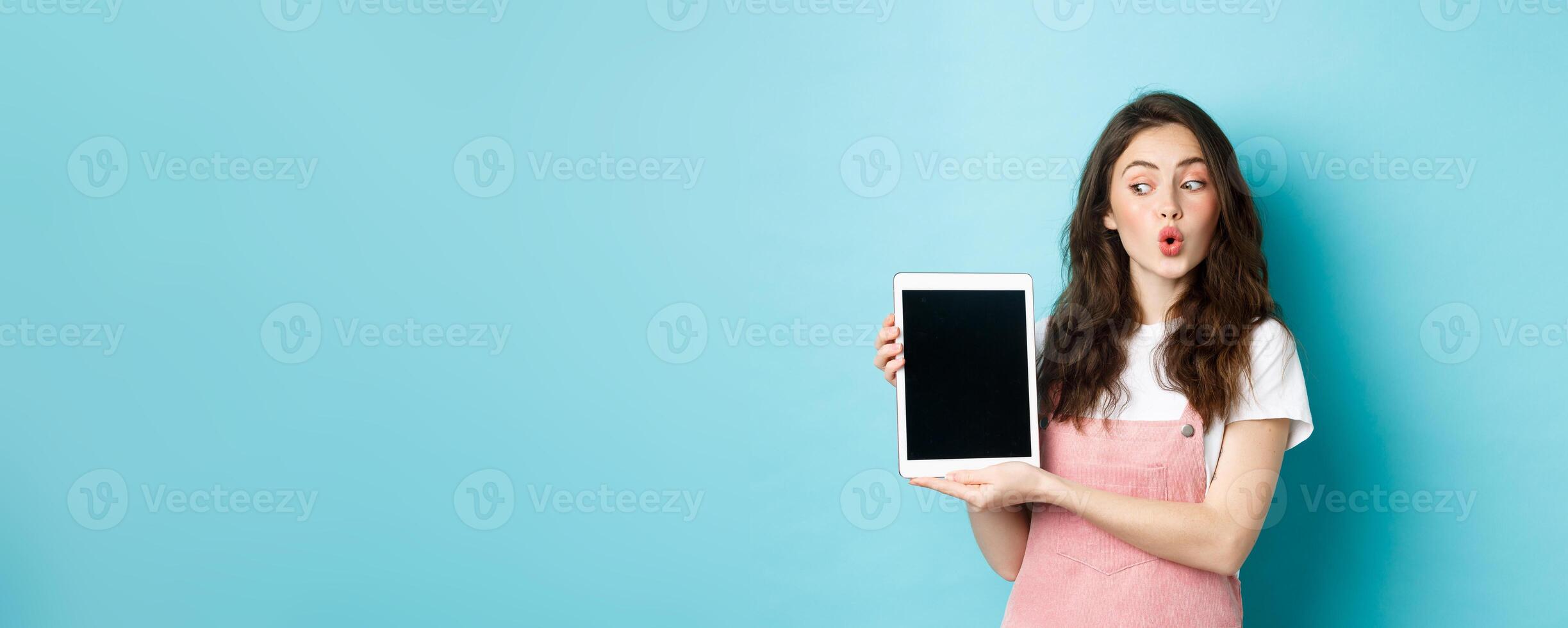 Intrigued glamour girl looking at digital tablet screen while showing it to you, standing amused with online promo offer, demonstrate something on display, blue background photo