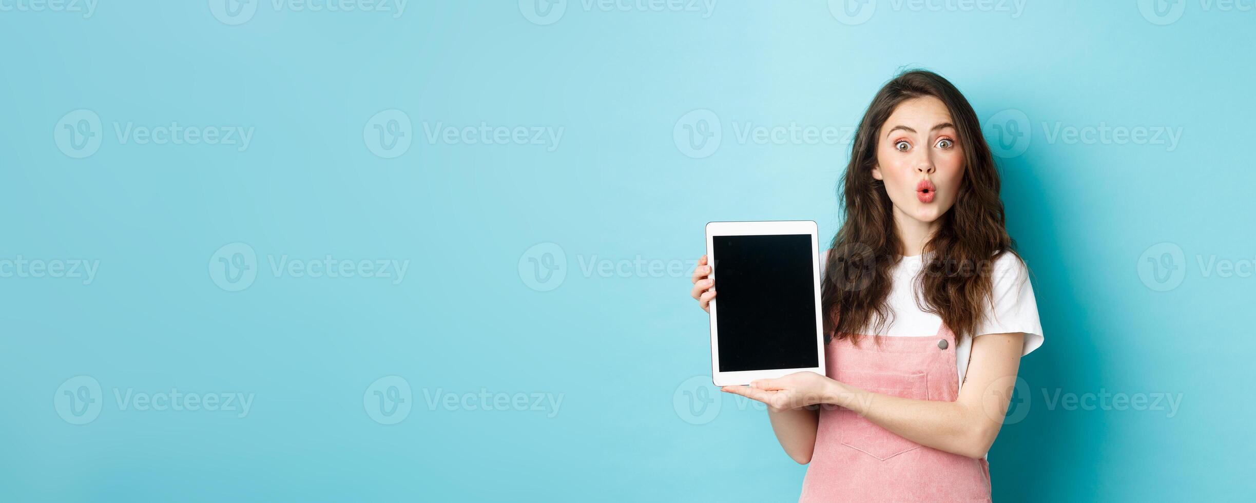 Surprised attractive female showing digital tablet screen and looking amazed at camera, checking out promo offer, standing amused against blue background photo