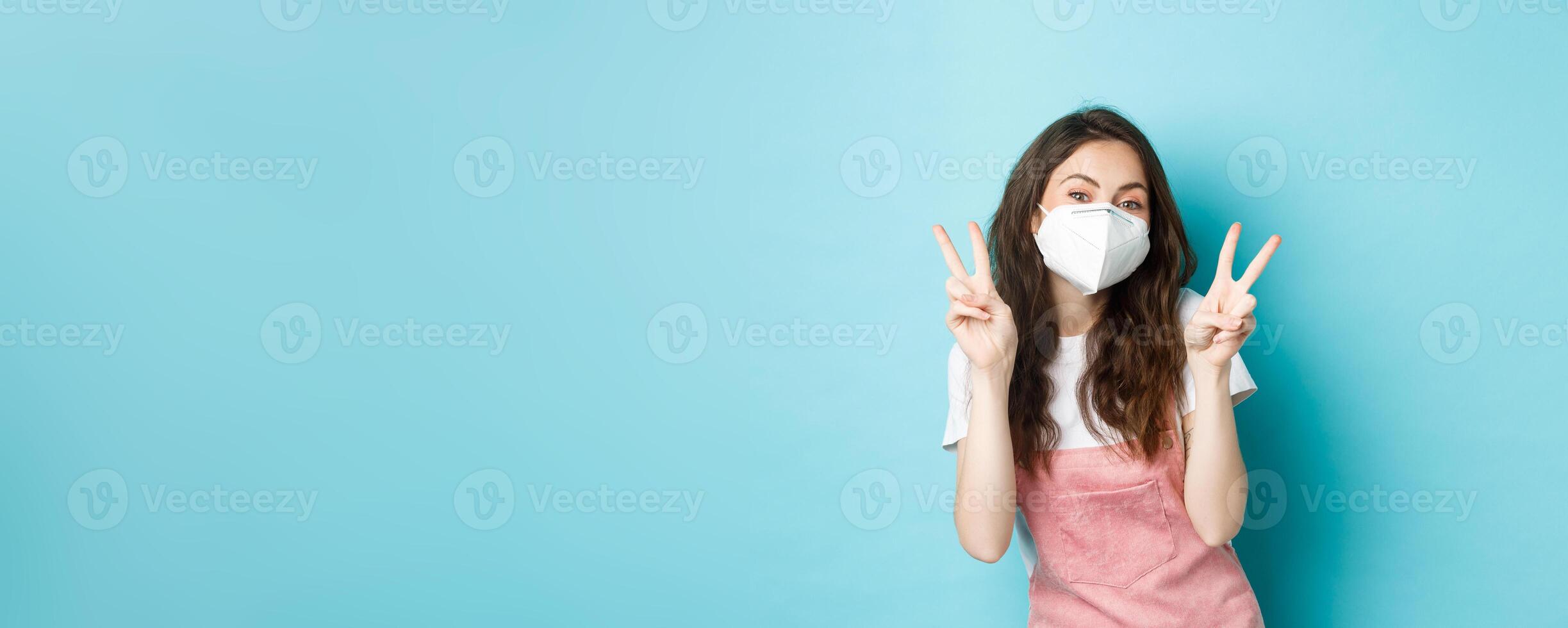 Health, coronavirus and social distancing concept. Happy young woman in medical respirator showing v-signs. Cute girl posing in face mask with peace gesture, blue background photo