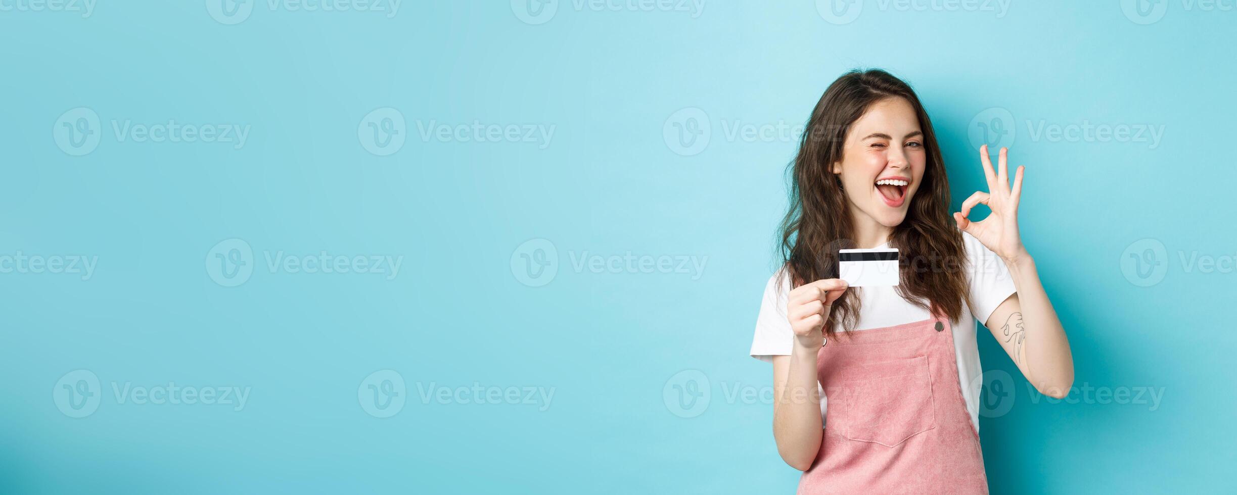 Cheerful brunette girl showing plastic credit card and okay sign, winking and smiling confident, recommend logo, standing over blue background photo